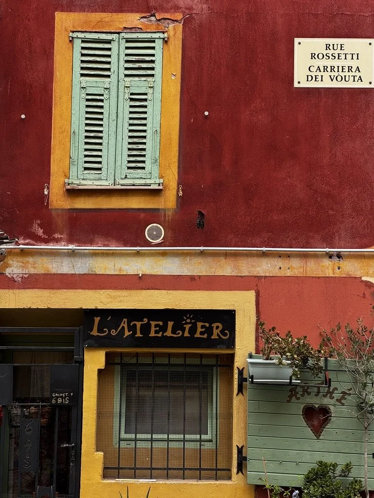 Colorful building with red upper wall, yellow window frame, and green window shutters; sign reading 'L'Atelier'; potted plants outside; street sign 'Rue Rosetti, Carriera Dei Vouta'.
