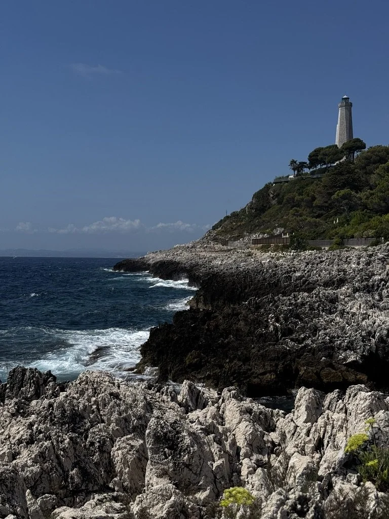 View of a rocky coastal shoreline with a lighthouse on a hill in the distance, under a blue sky.