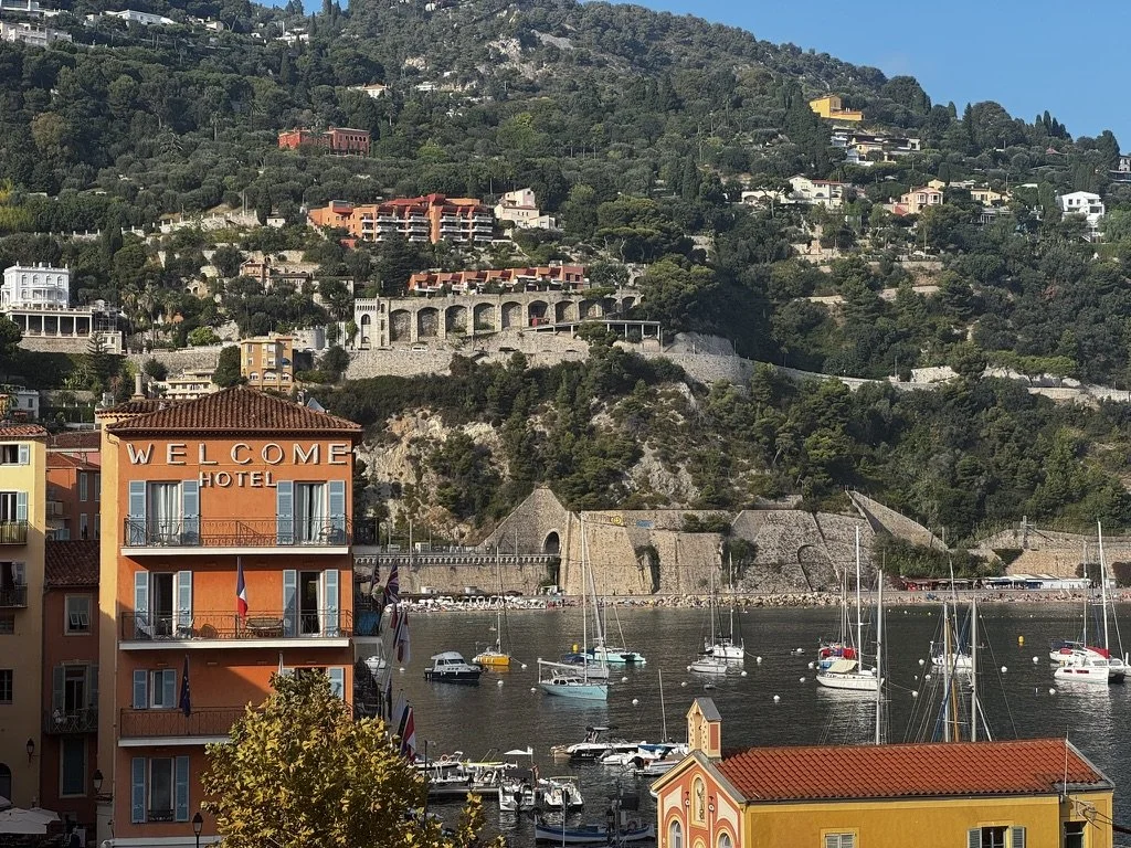 A scenic harbor with sailboats docked, colorful buildings including a prominent orange hotel with a welcome sign, and a hillside with lush greenery and residential structures in the background.