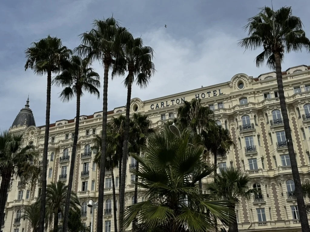 The Carlton Hotel building with palm trees in front and a cloudy sky overhead. French Riviera, Canne, France