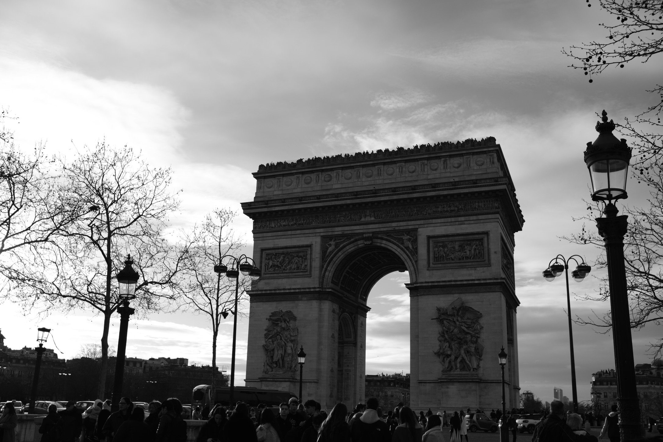 Black and white photo of the Arc de Triomphe in Paris with people and street lamps in the foreground, bare trees, and a cloudy sky.