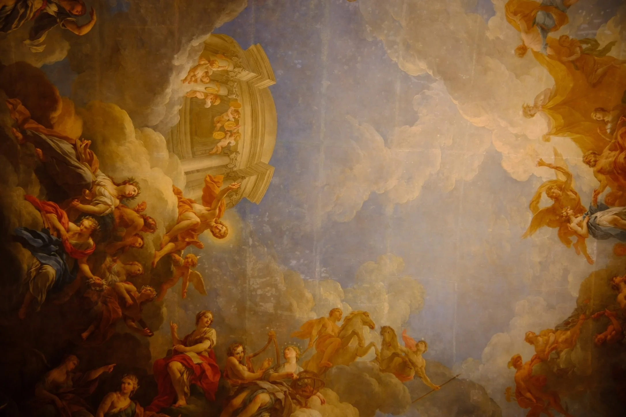 Baroque-style ceiling painting depicting mythological figures, clouds, and angels in the sky with a classical building in the background. Shot in Palace of Versailles, France