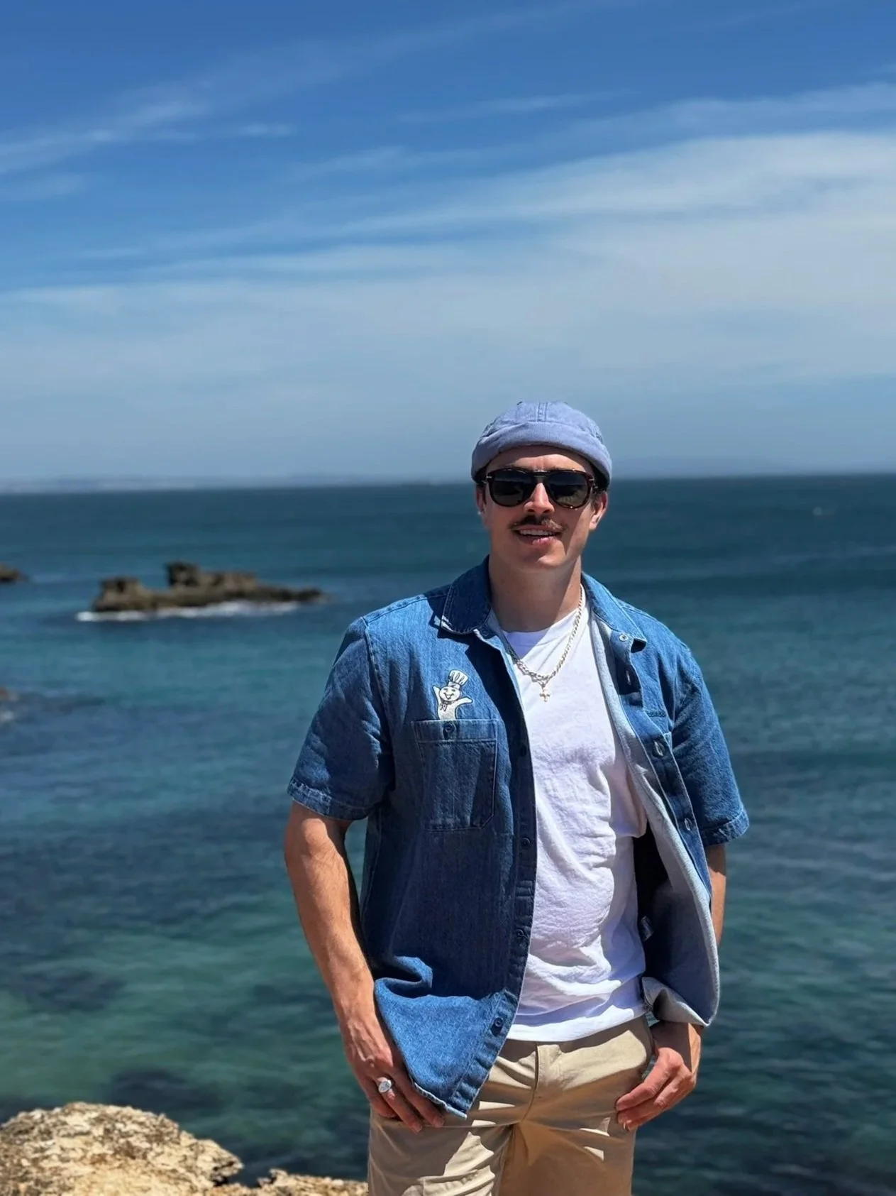 A man with a mustache wearing sunglasses, a gray beanie, a white t-shirt, and a denim jacket standing by the ocean with rocks and blue sky in the background.