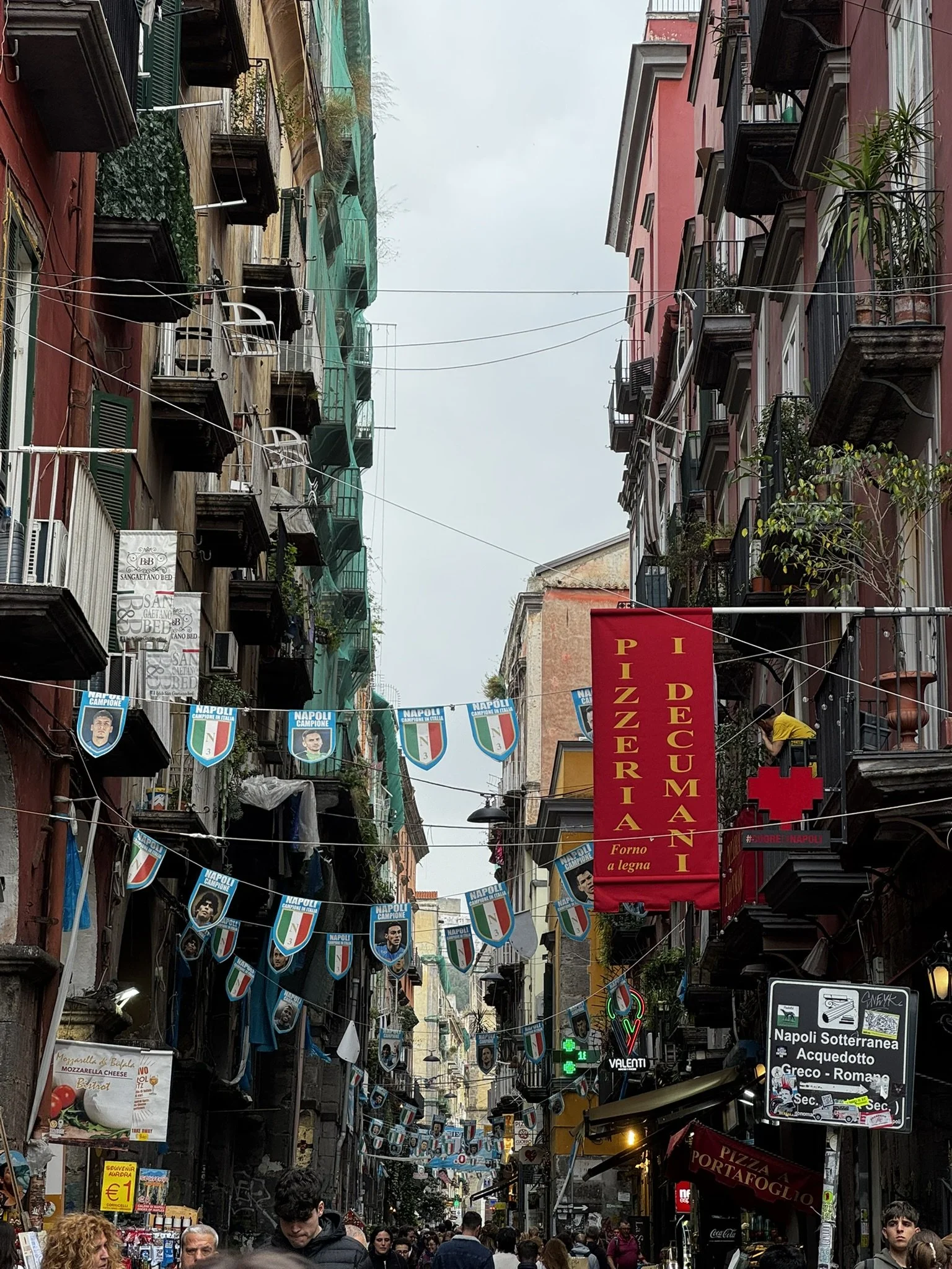 City street filled with pedestrians, decorated with banners and flags supporting Napoli football team, several businesses including a pizzeria, pharmacy, and food stalls, with tall, narrow buildings typical of a European city.