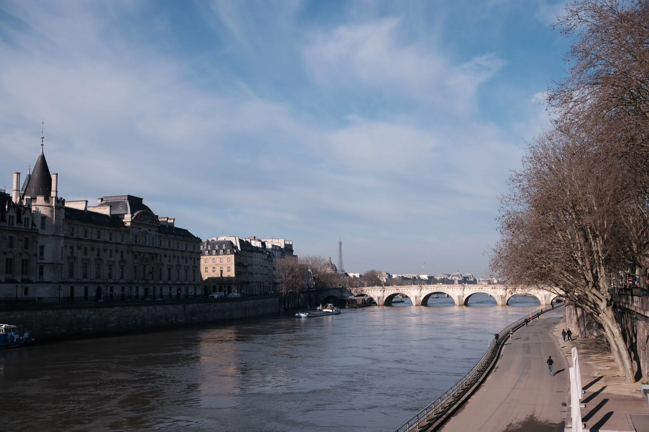 View of the Seine River in Paris with historic buildings on the left, a stone bridge in the background, bare trees on the right, and the Eiffel Tower visible in the distance under a partly cloudy sky.
