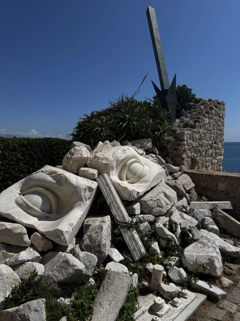 Sculpture of two faces carved into white stone blocks, with metal and stone elements around and above it, located outdoors near a stone wall and overlooking the ocean.