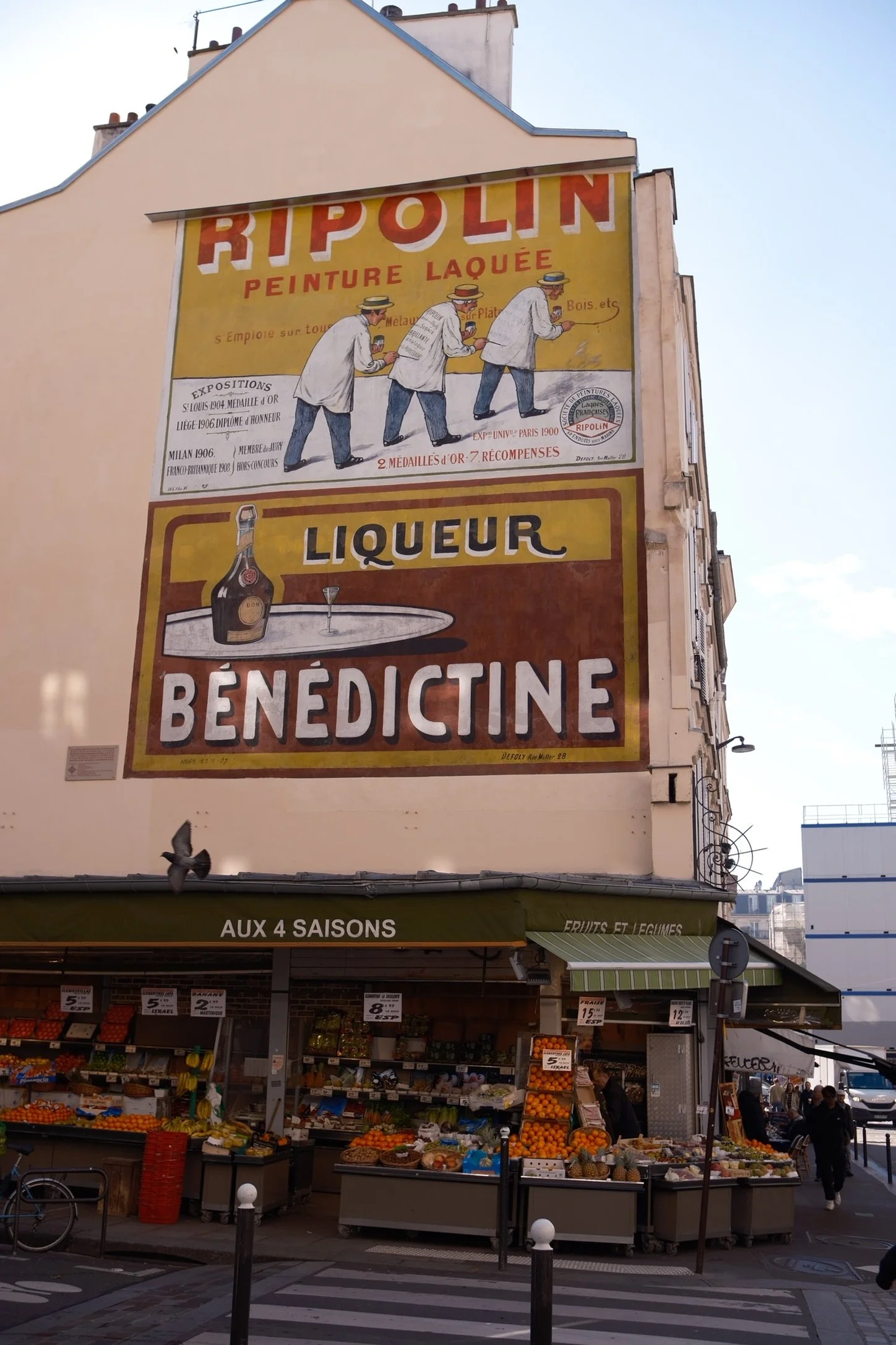 A street view with a fruit and vegetable stand in front of a building. The building has large vintage-style advertisements, including one for a paint company called Ripolin, featuring men painting. A smaller sign below advertises a liqueur named Bene