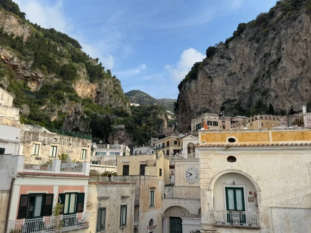 View of a town with colorful buildings nestled against rocky cliffs and green hillside under a blue sky. Shot in Amalfi Coast, Italy