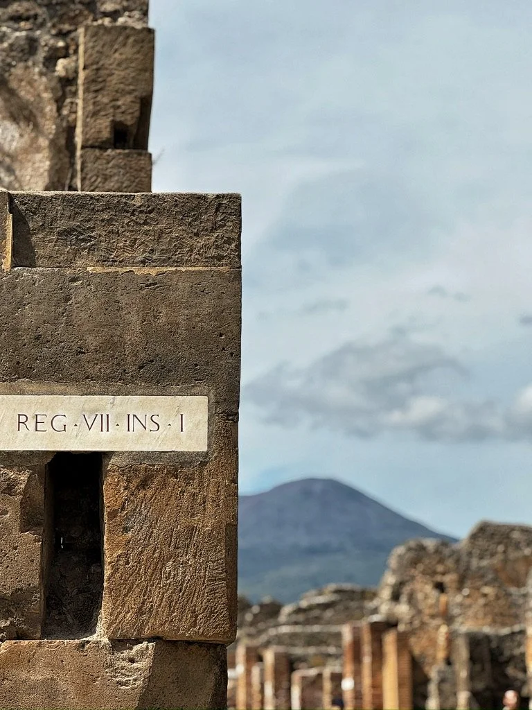 Close-up of ancient stone ruins with an inscribed plaque reading "REG·VII·INS·I," and a mountain in the background under a partly cloudy sky. Shot in Pompeii, Italy