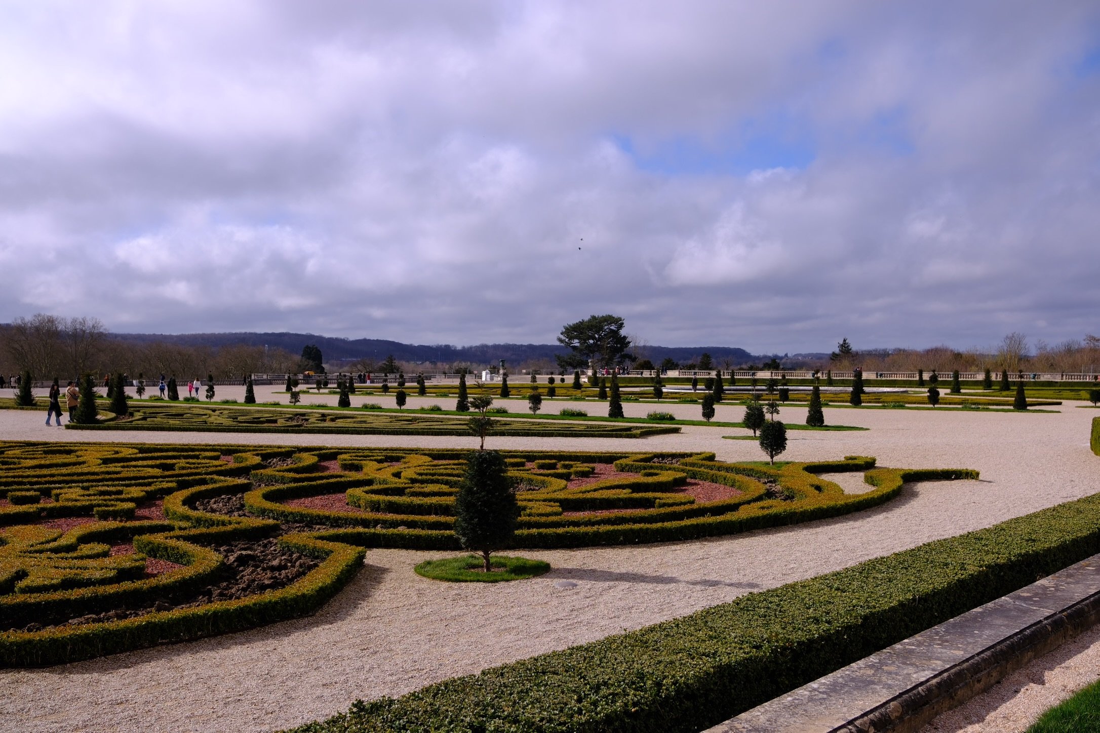 View of a formal garden with intricate map-like hedge designs, small trees, and walkways against a cloudy sky and distant hills. Shot in Palace of Versailles, France 
