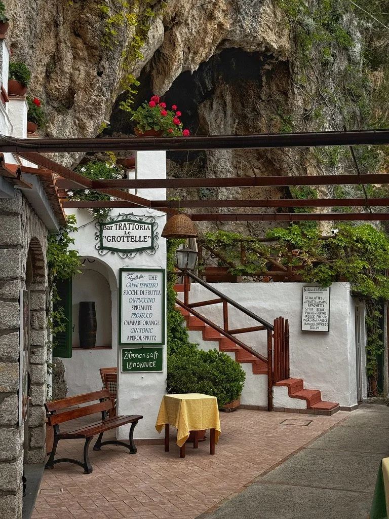 Outdoor seating area of a restaurant named 'Le Grotteelle' with a yellow tablecloth-covered table, benches, and stairs with greenery and flowers, built into a rocky cliffside. Shot in Capri, Italy