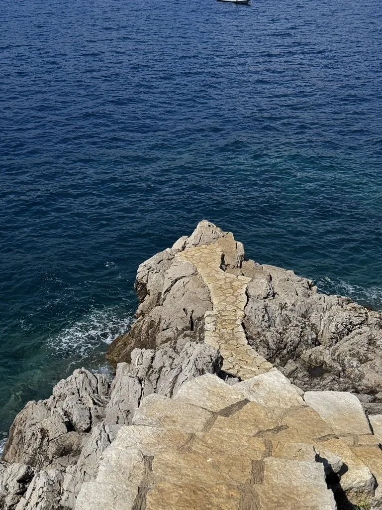 A stone pathway extending over rocky coastline into the ocean, with blue water beneath and a small boat visible in the distance.