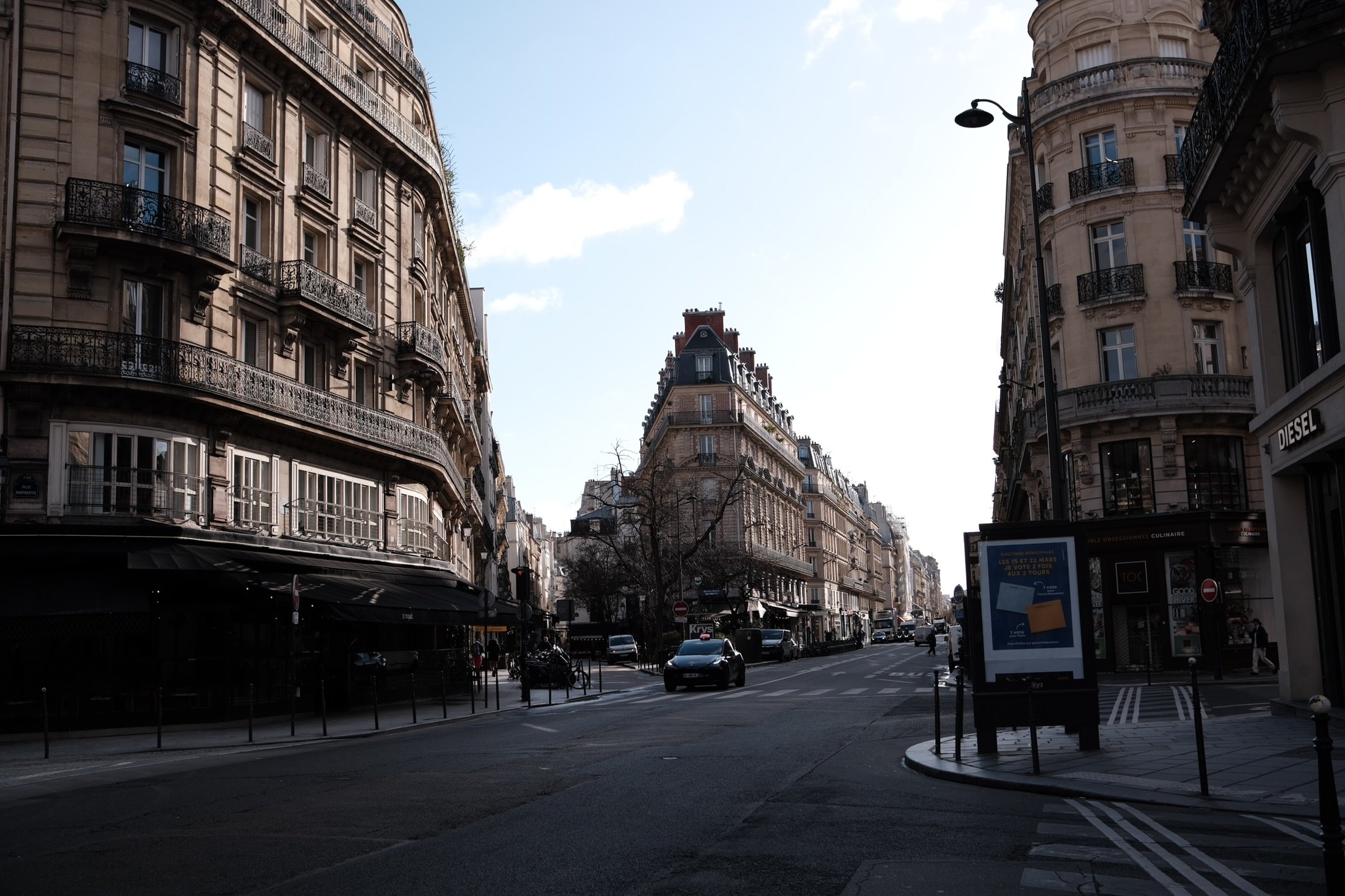 Paris city street with classic European architecture, cars on the road, a large digital billboard, and a clear sky.