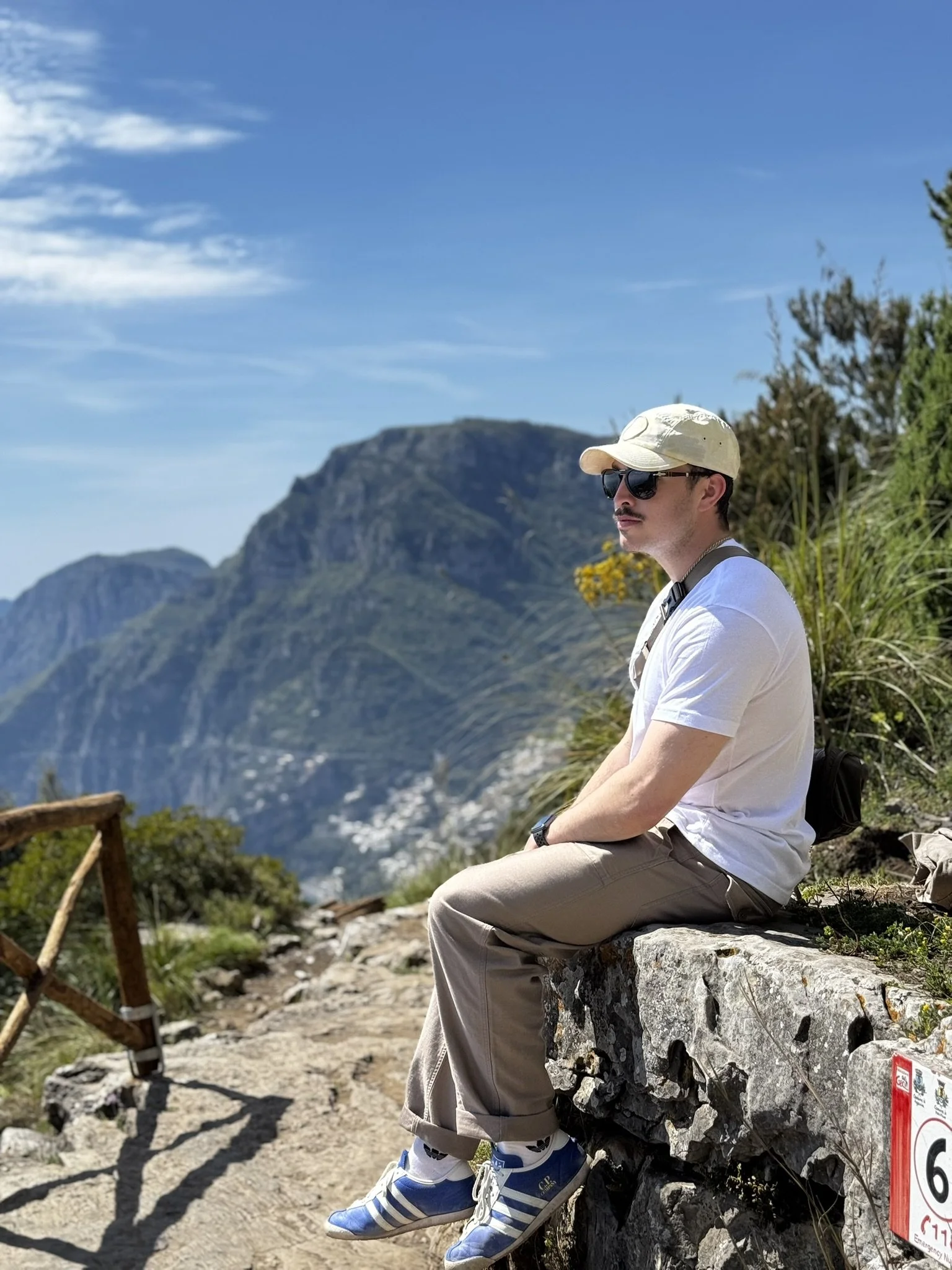 A man with sunglasses, a cap, white t-shirt, beige pants, and blue sneakers sitting on a rock ledge during a mountain hike with a scenic mountain landscape in the background. Path of the Gods, Amalfi Coast, Italy