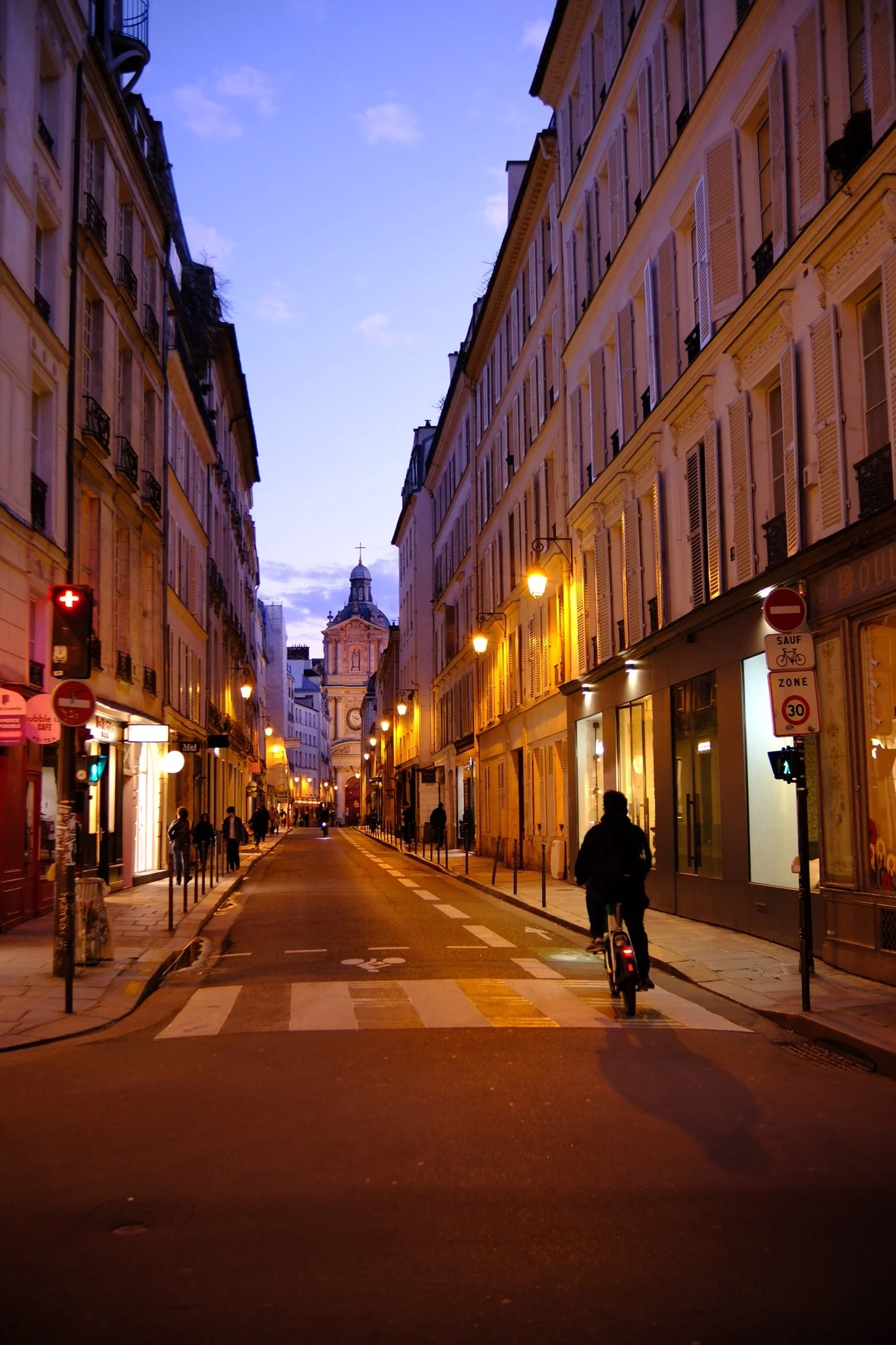 Paris street scene during evening with tall buildings, streetlights, pedestrians, and a cyclist riding down a narrow road leading to a church in the distance.