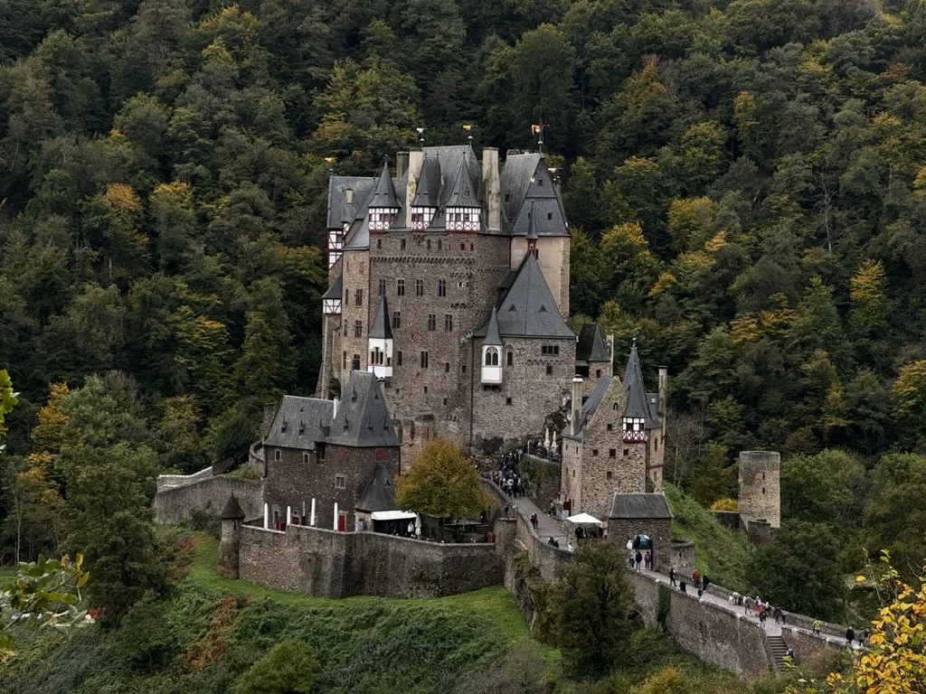 A medieval castle surrounded by a forested hillside with autumn-colored trees, featuring tall towers with pointed roofs and stone walls. Berg-Etz Castle, Germany