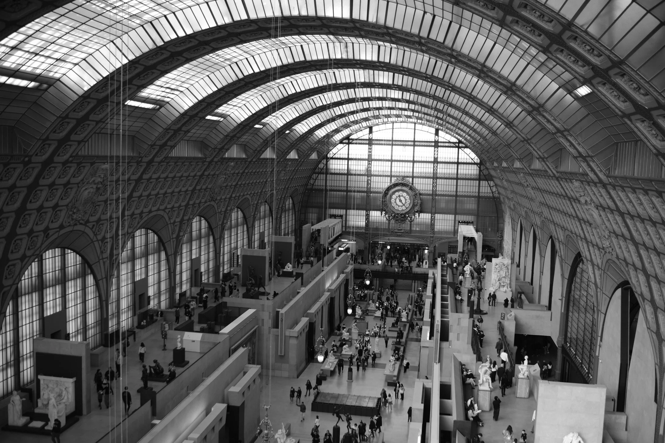 Interior of a grand historic Museum d'Orsay in Paris. Picture includes arched glass ceiling, large clock, and multiple levels of people and galleries.