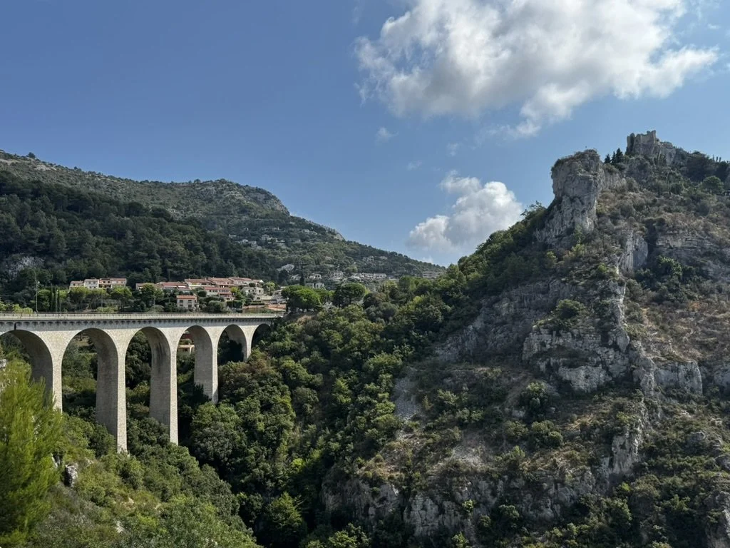 A stone arch bridge crossing a lush green valley with rocky hills and a small town in the background under a partly cloudy sky. Eze, French Riviera, France