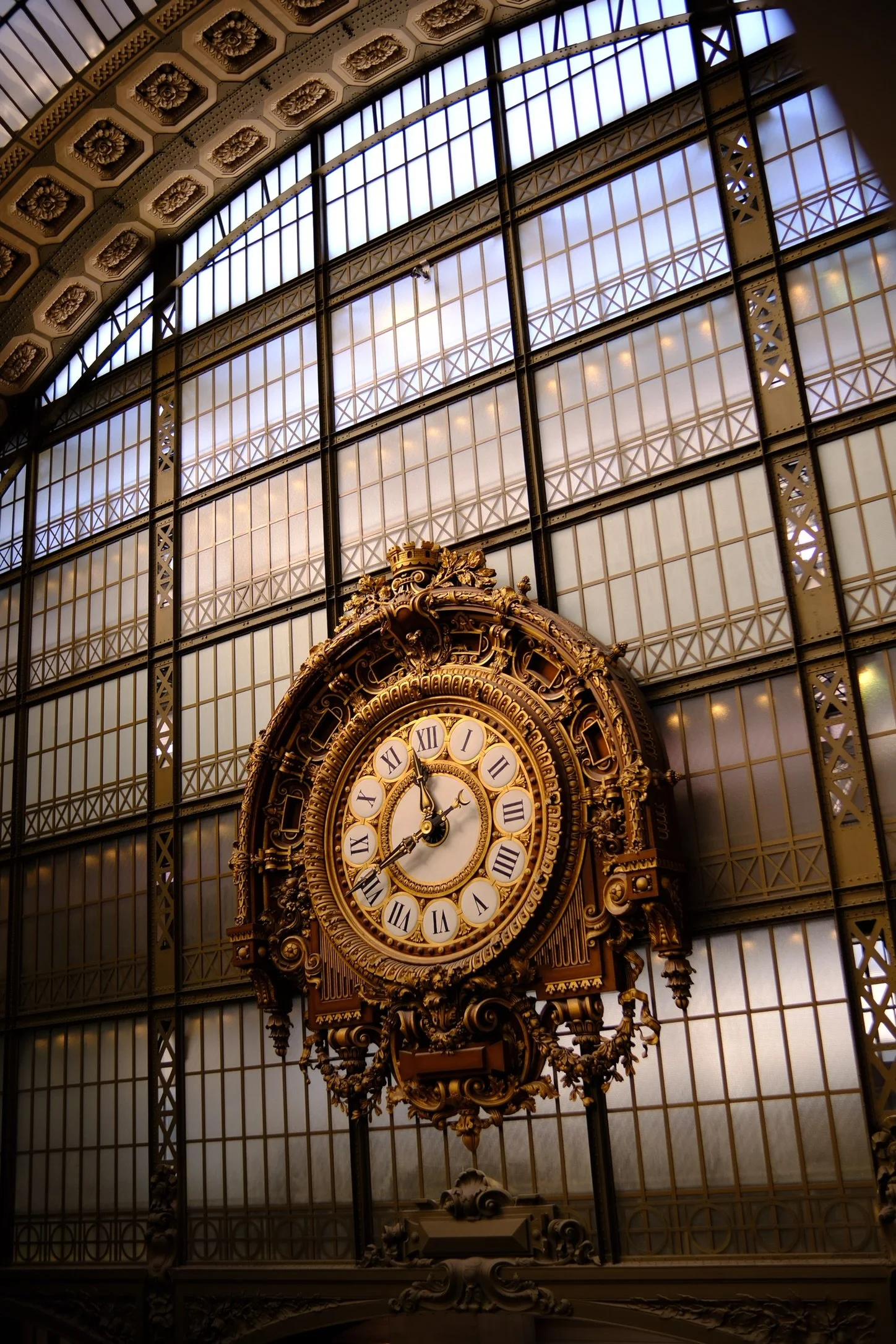 An ornate, antique clock in the Museum d'Orsay with Roman numerals and gold detailing inside a glass-ceilinged building.