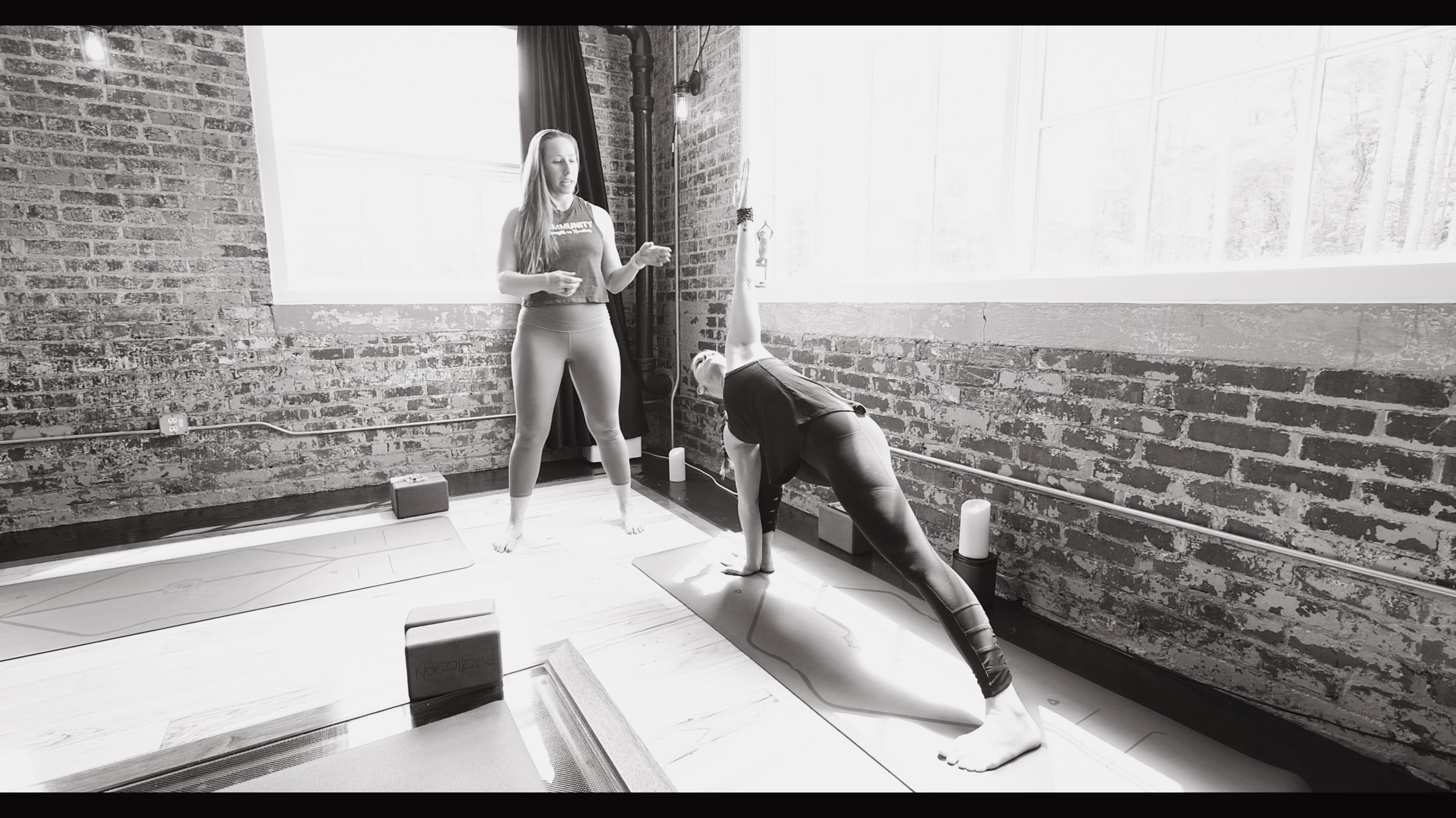 A woman practicing yoga in a studio with exposed brick walls, sunlight streaming through large windows, and a yoga instructor nearby.