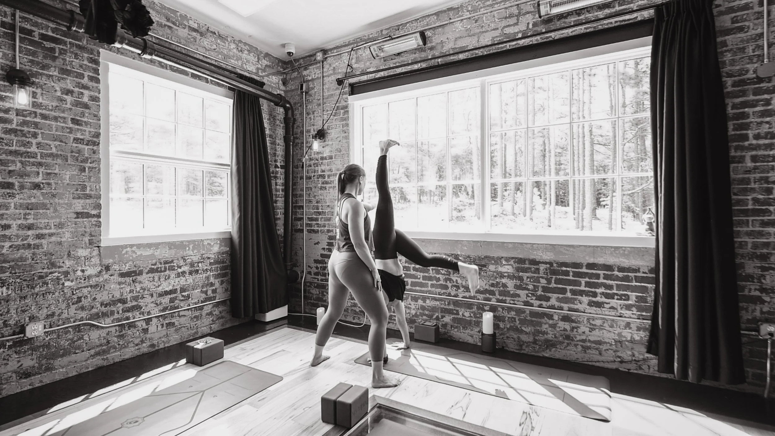 Two women practicing yoga in a studio with exposed brick walls and large windows, one assisting the other with a handstand.