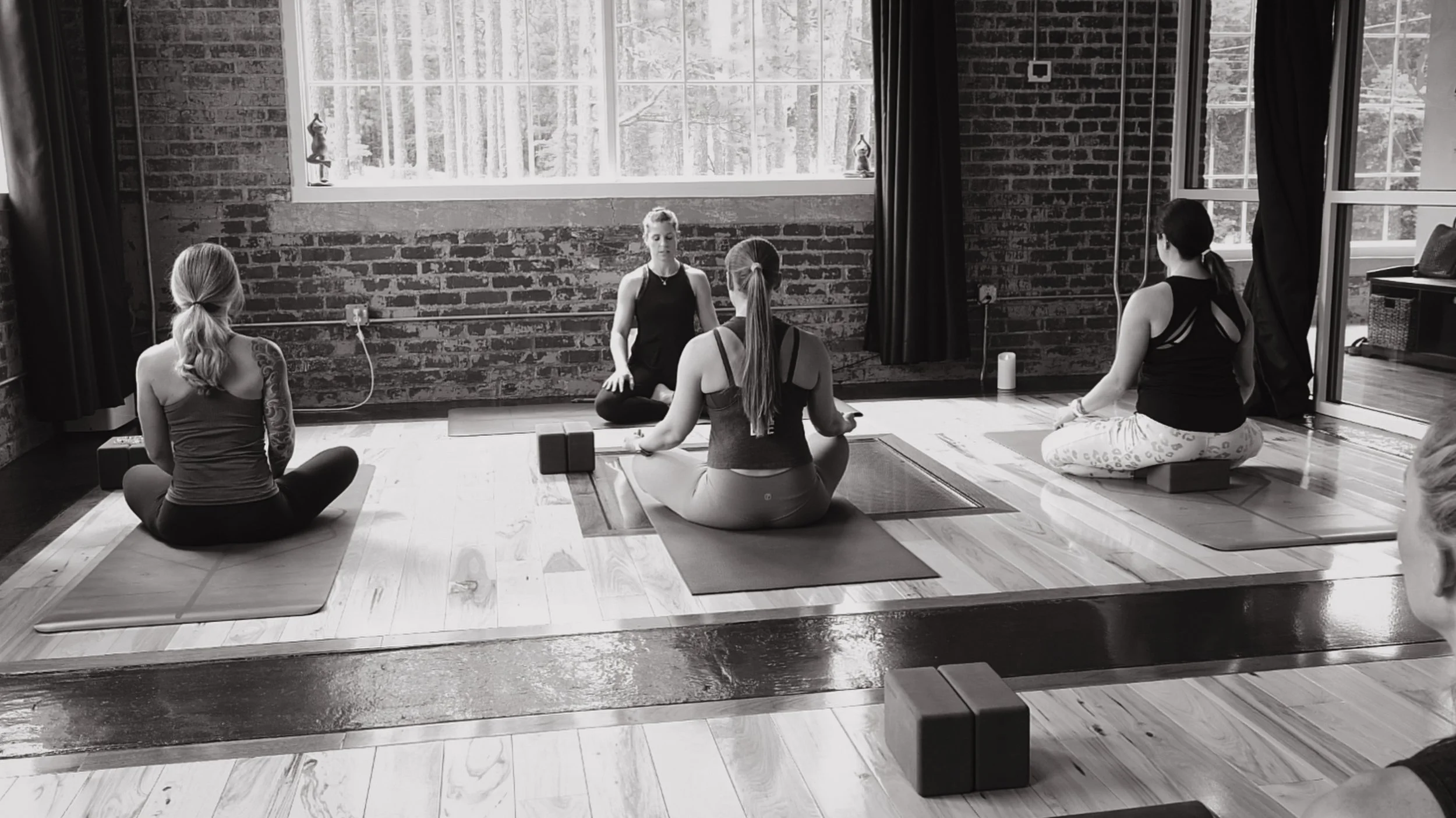 Four women participating in a yoga class in a studio with brick walls and large windows, sitting on yoga mats in a cross-legged position.