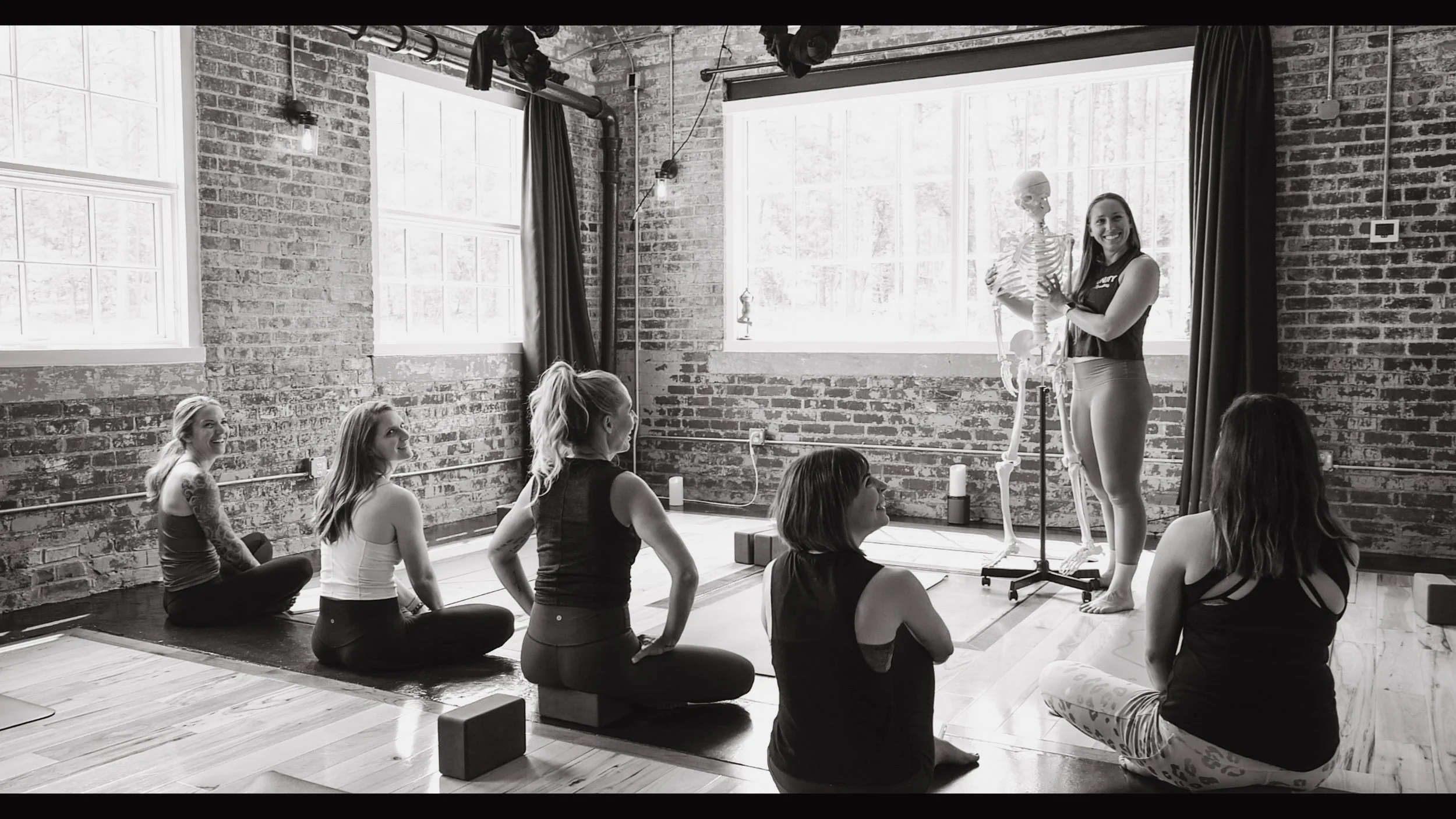 A yoga instructor holding a skeleton model stands in front of five women seated on yoga mats, in a brick-walled studio with large windows, during a yoga class.