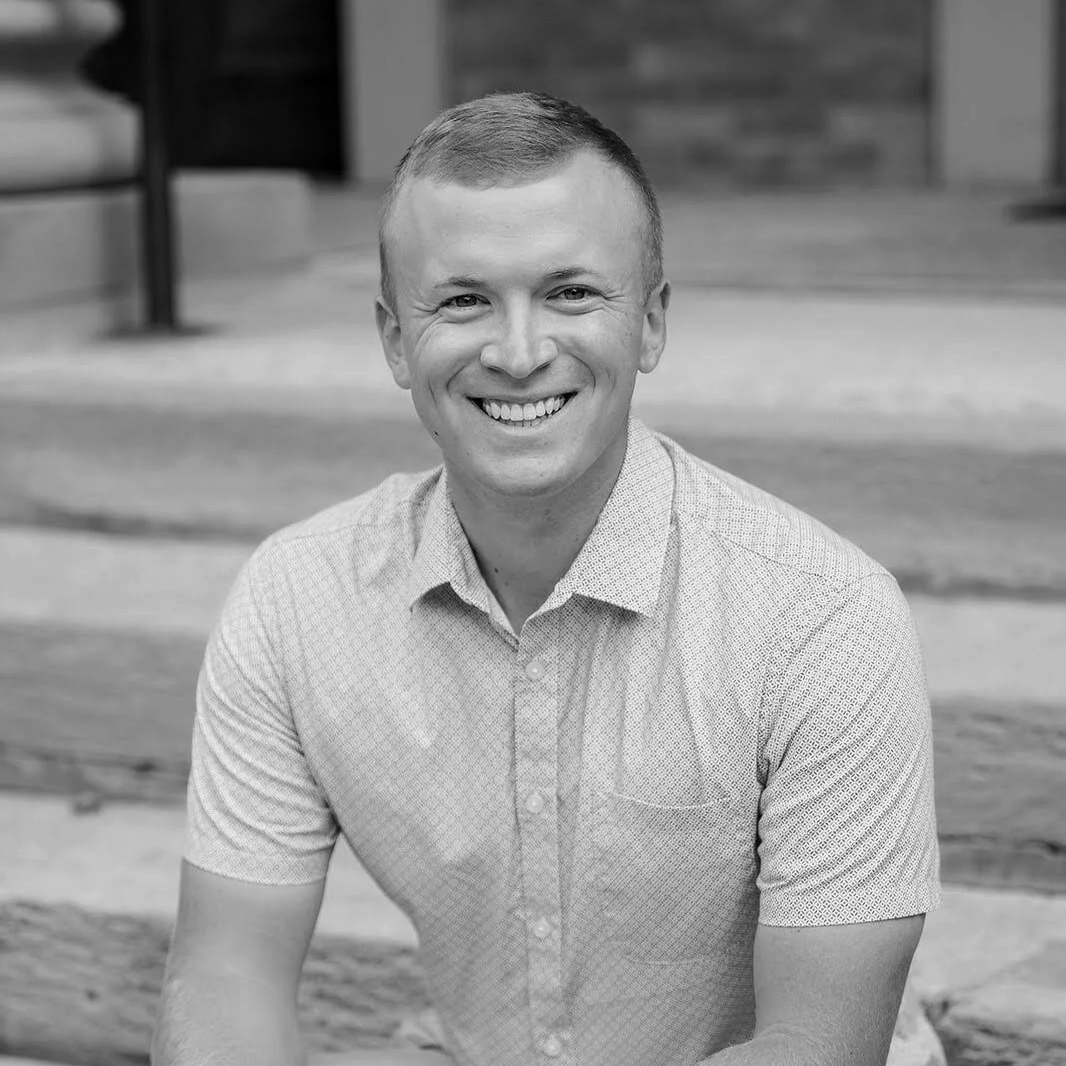 Taylor Suggs, smiling, wearing a light-colored, short-sleeved button-up shirt, sitting outdoors.