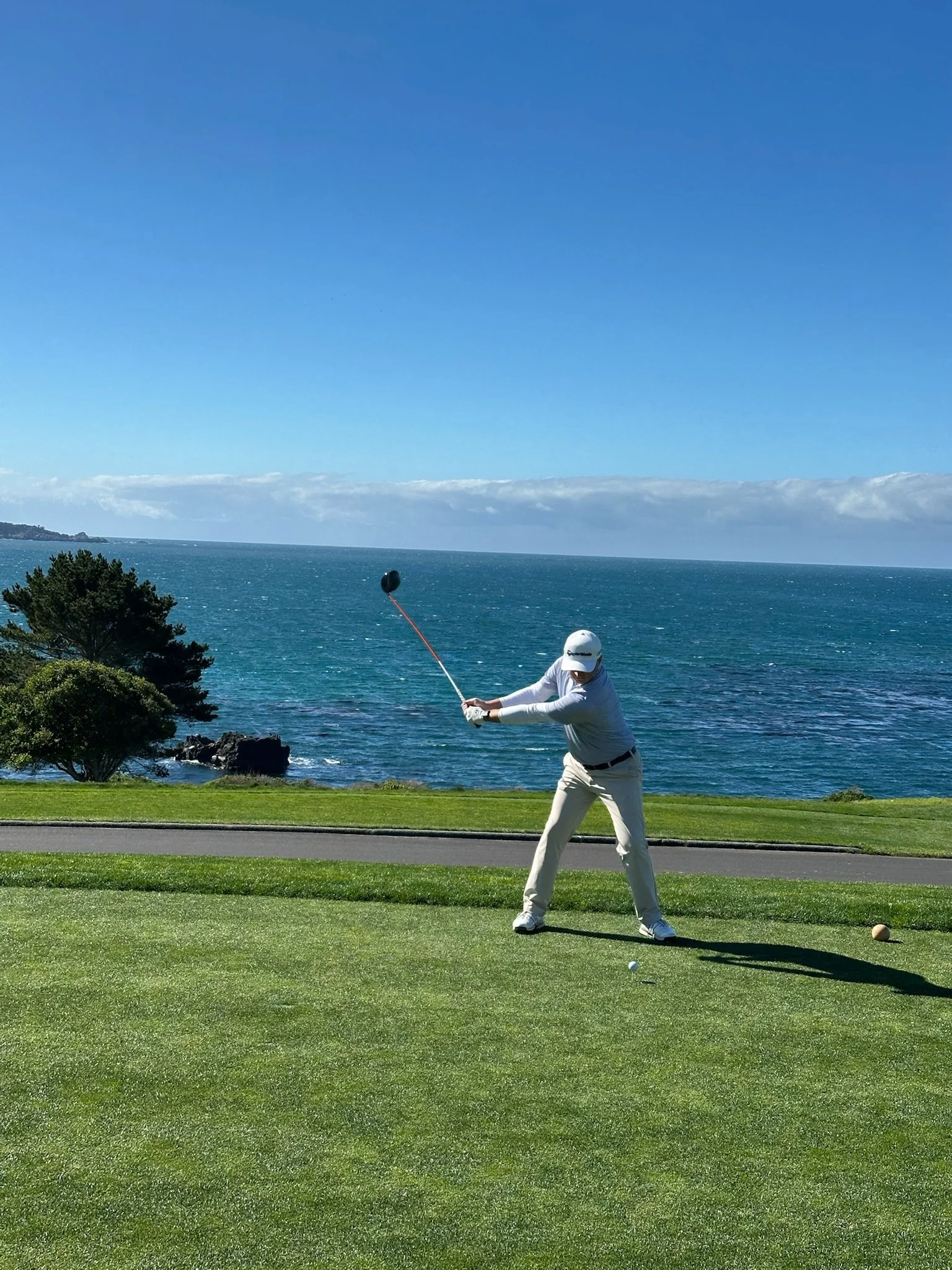 Aerial view of a coastline with rocks in the ocean, waves crashing against them, and a green golf course with sand traps and pathways on the land.