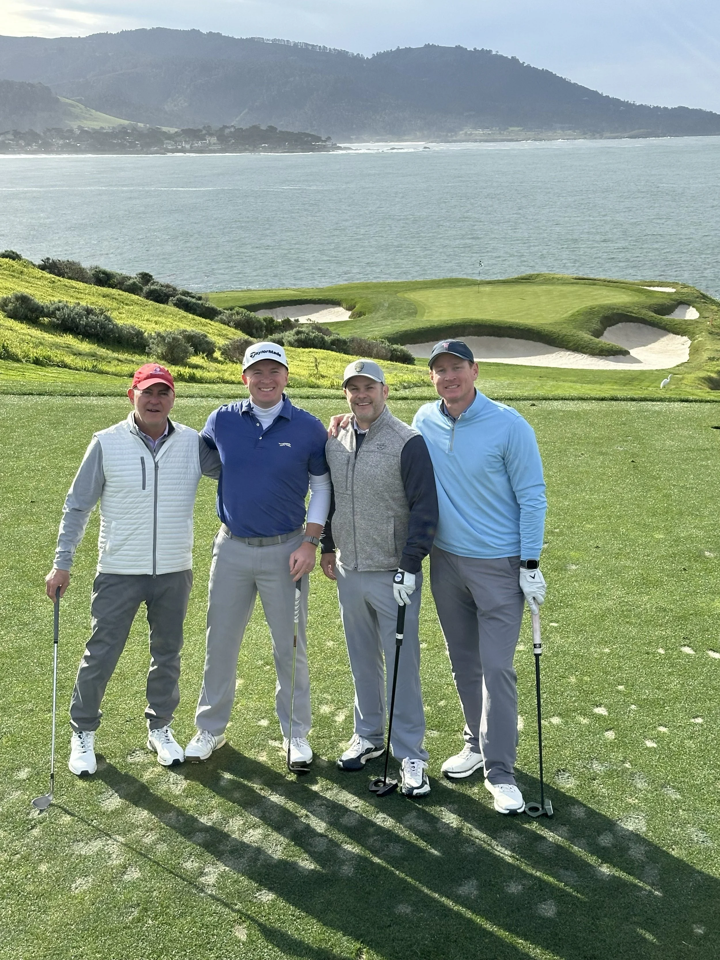 Coastal scene with a golf hole and ocean under a clear blue sky, with distant mountains on the horizon.