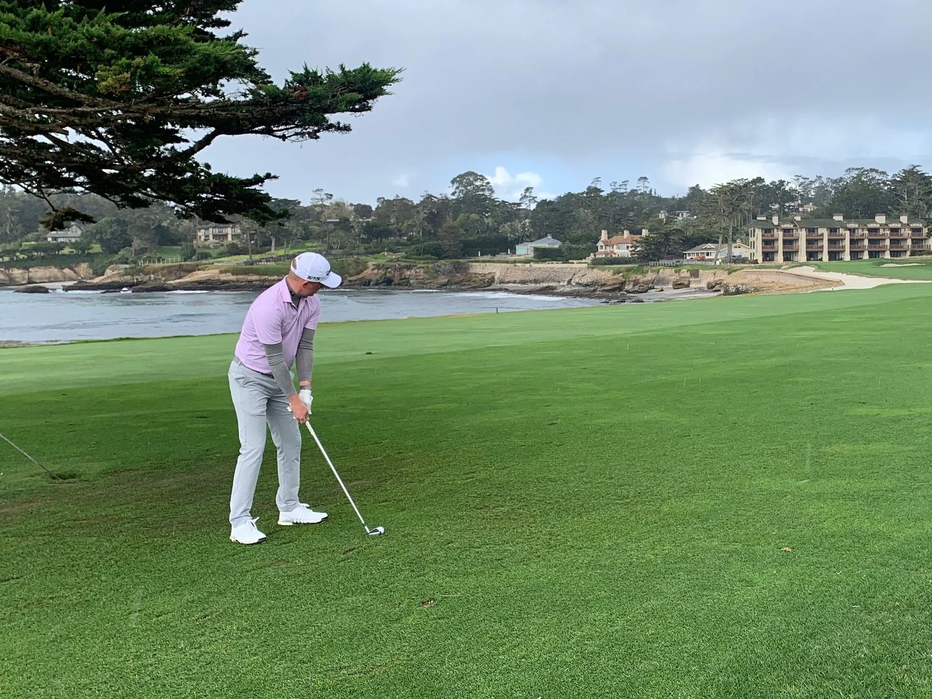 A golf course near the ocean with a green, flag, and sand trap, along with buildings and cliffs in the background under a cloudy sky.