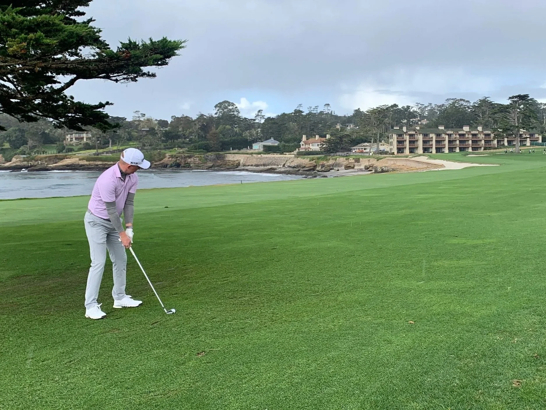 A golf course near the coast with a green, a flag, and a sand bunker in the foreground, a cliff and ocean in the background, and buildings and facilities on the hilltop under a blue sky.