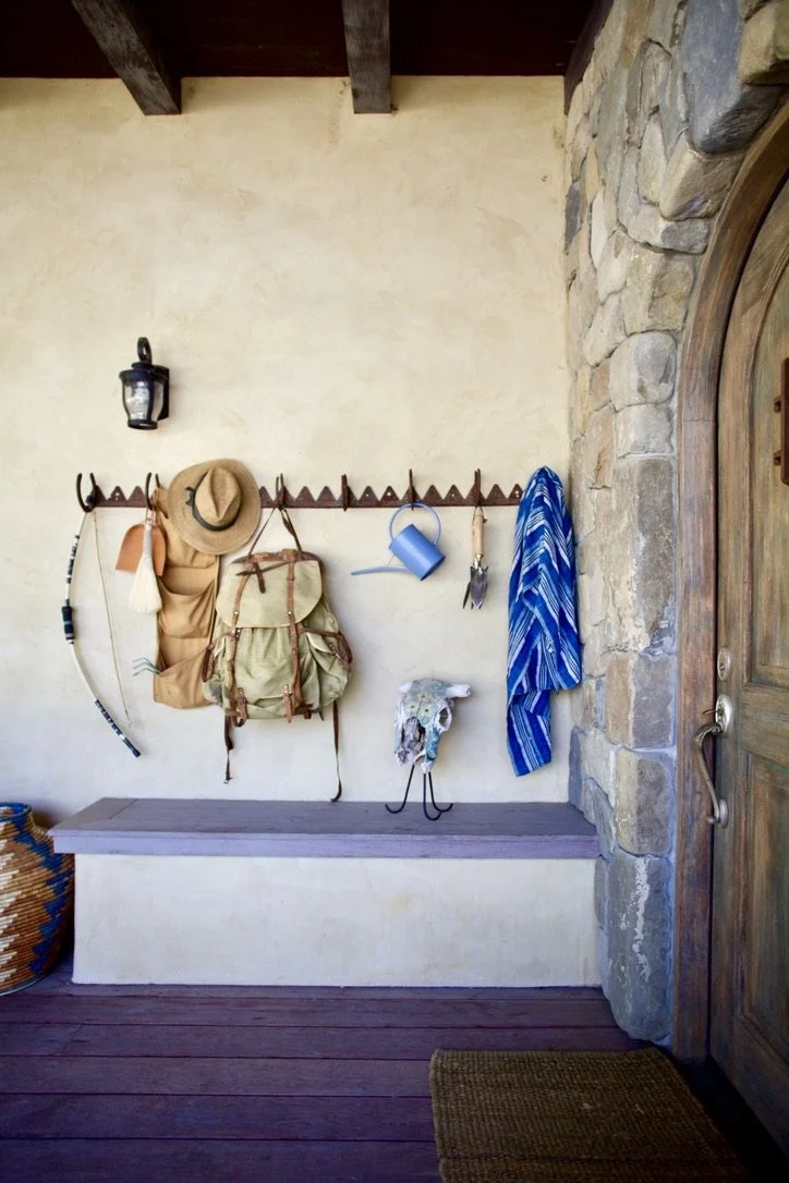 Outdoor entryway with hooks on a wall holding a backpack, straw hat, towel, blue jug, and a scarf. There is a bench below and a stone wall next to a wooden door.