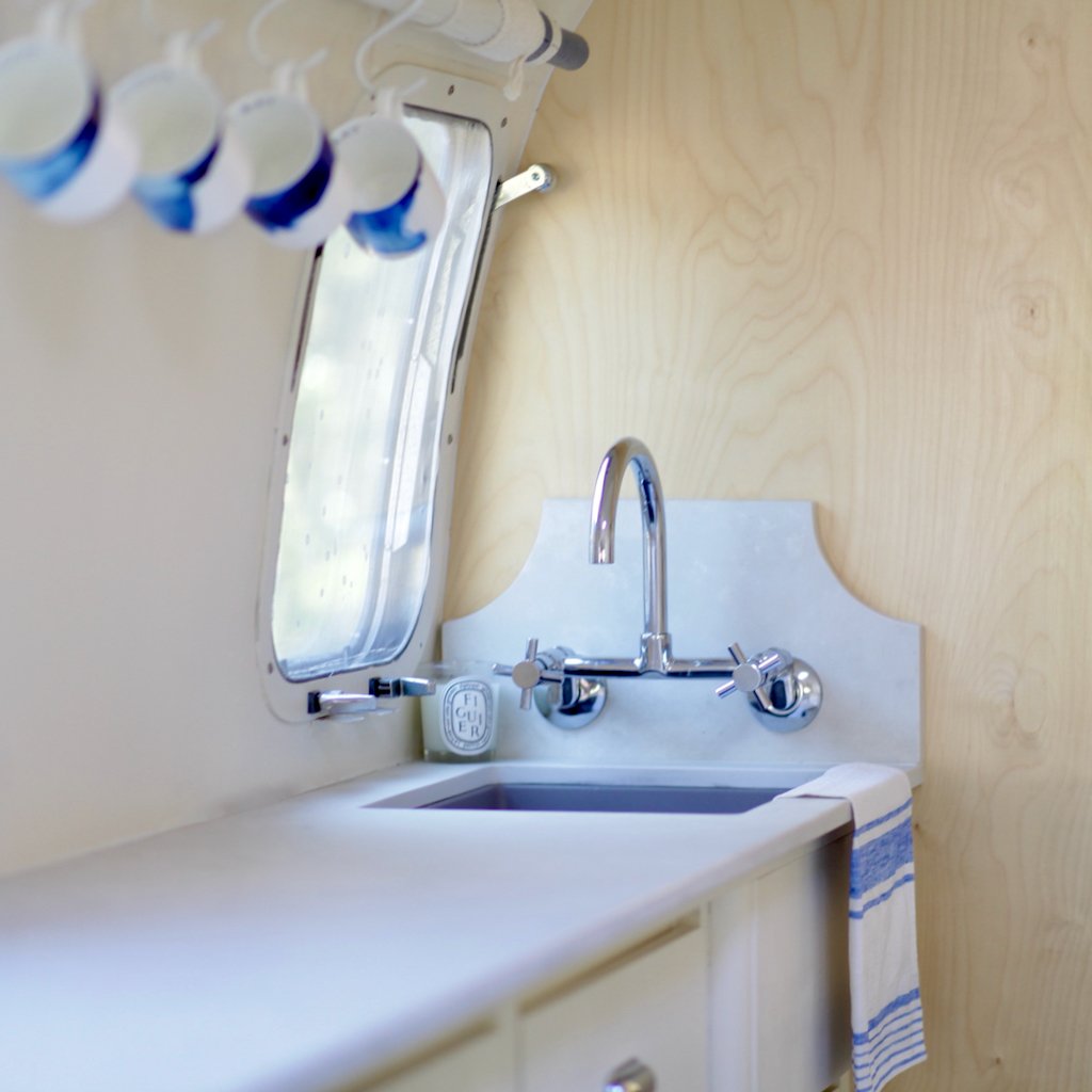 Small kitchen area with a stainless steel faucet and sink, a white countertop, a striped towel hanging, and a window with a curved frame and upper shelf holding part of a row of small bottles.