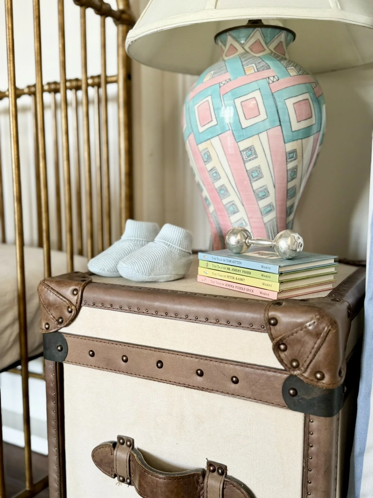 Close-up of a vintage-style suitcase used as a side table, with a stack of children's books, a silver dumbbell, and a pair of white baby socks on top. In the background, a colorful ceramic table lamp with a geometric pattern.