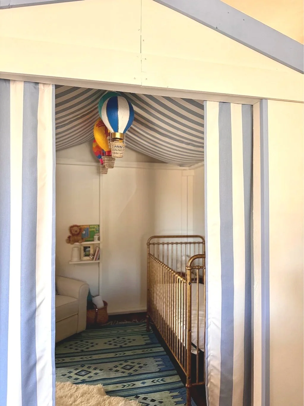 Interior of a cozy nursery with hot air balloon decorations hanging from a blue and white striped ceiling canopy, a gold crib on the right, a small shelving unit with books and a plush teddy bear on the left, and a colorful rug on the floor.