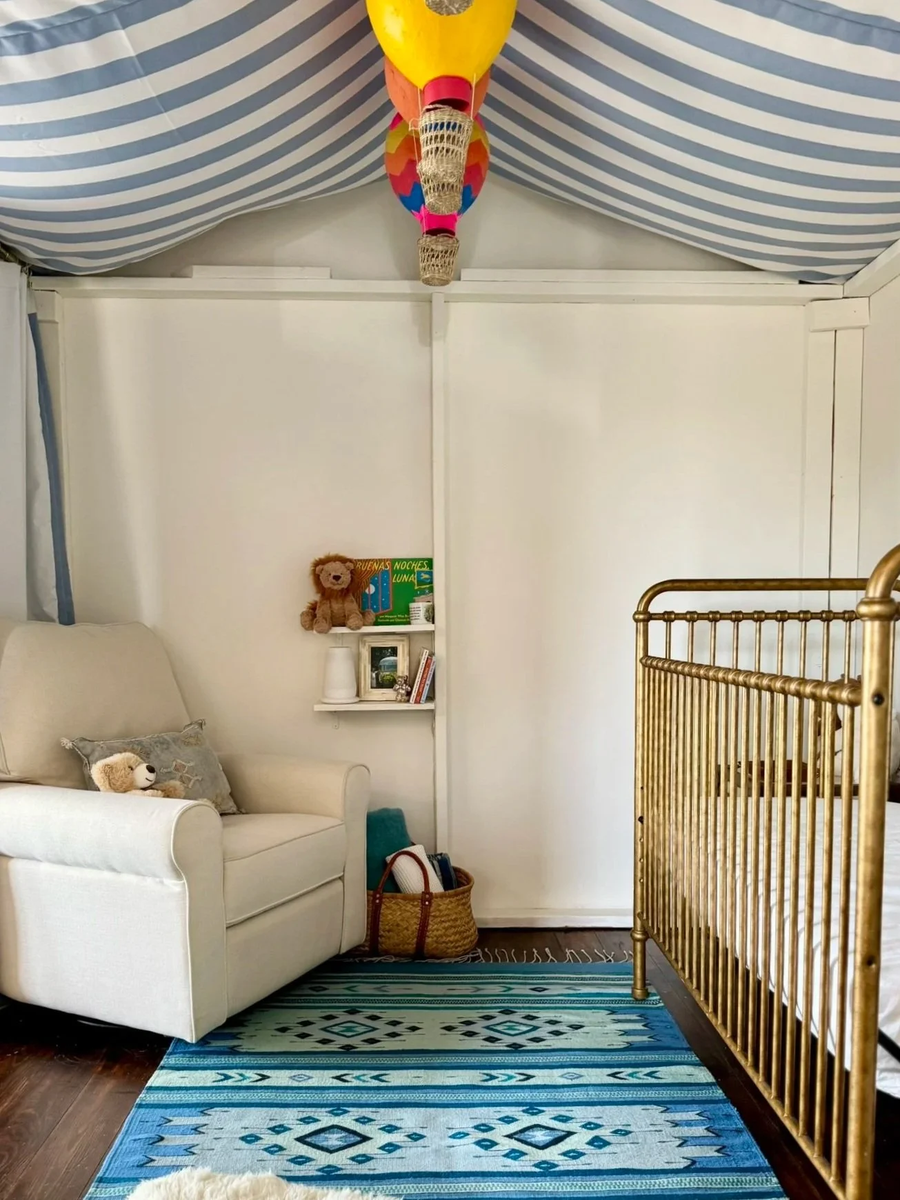 A cozy nursery corner with a white armchair, a small pillow, a teddy bear, a basket with books and toys, and a golden crib on a patterned blue rug. The wall has floating shelves with stuffed animals and framed pictures, and colorful hot air balloon d