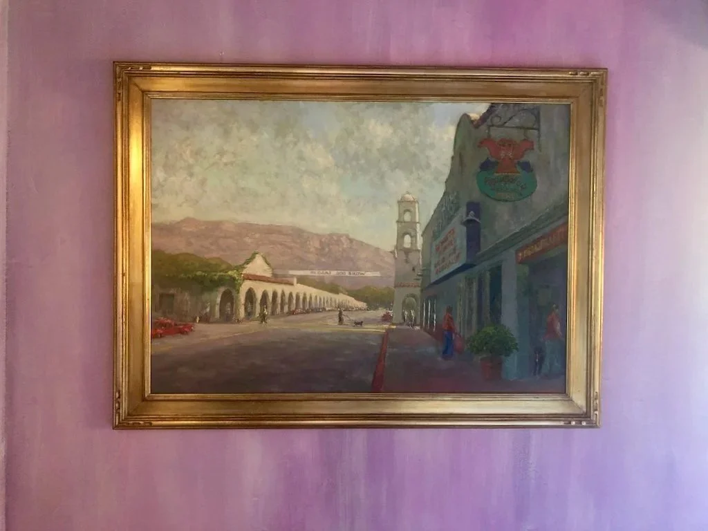 A framed landscape oil painting depicting a street scene with building arches and a clock tower, set against distant mountains and sky, hung on a purple wall.