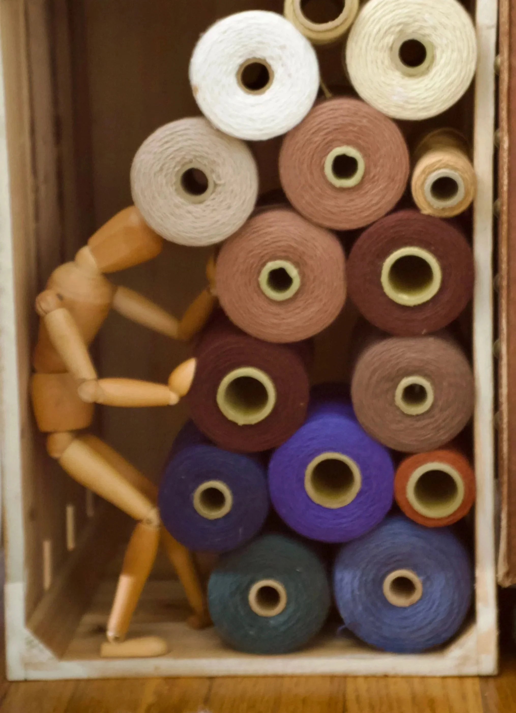 A wooden artist's mannequin sitting on the edge of a box filled with yarn spools in various colors, inside a wooden box.
