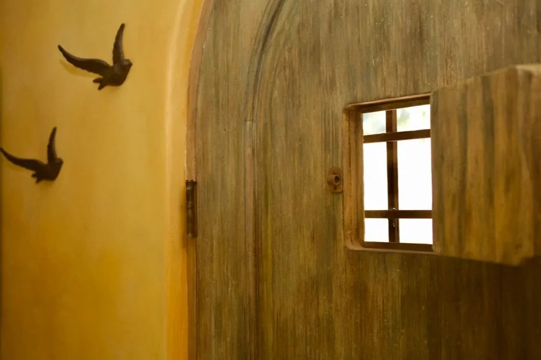 Close-up of a small wooden window with a grid design on a textured yellow wall. Two black bird-shaped wall decorations are visible nearby.