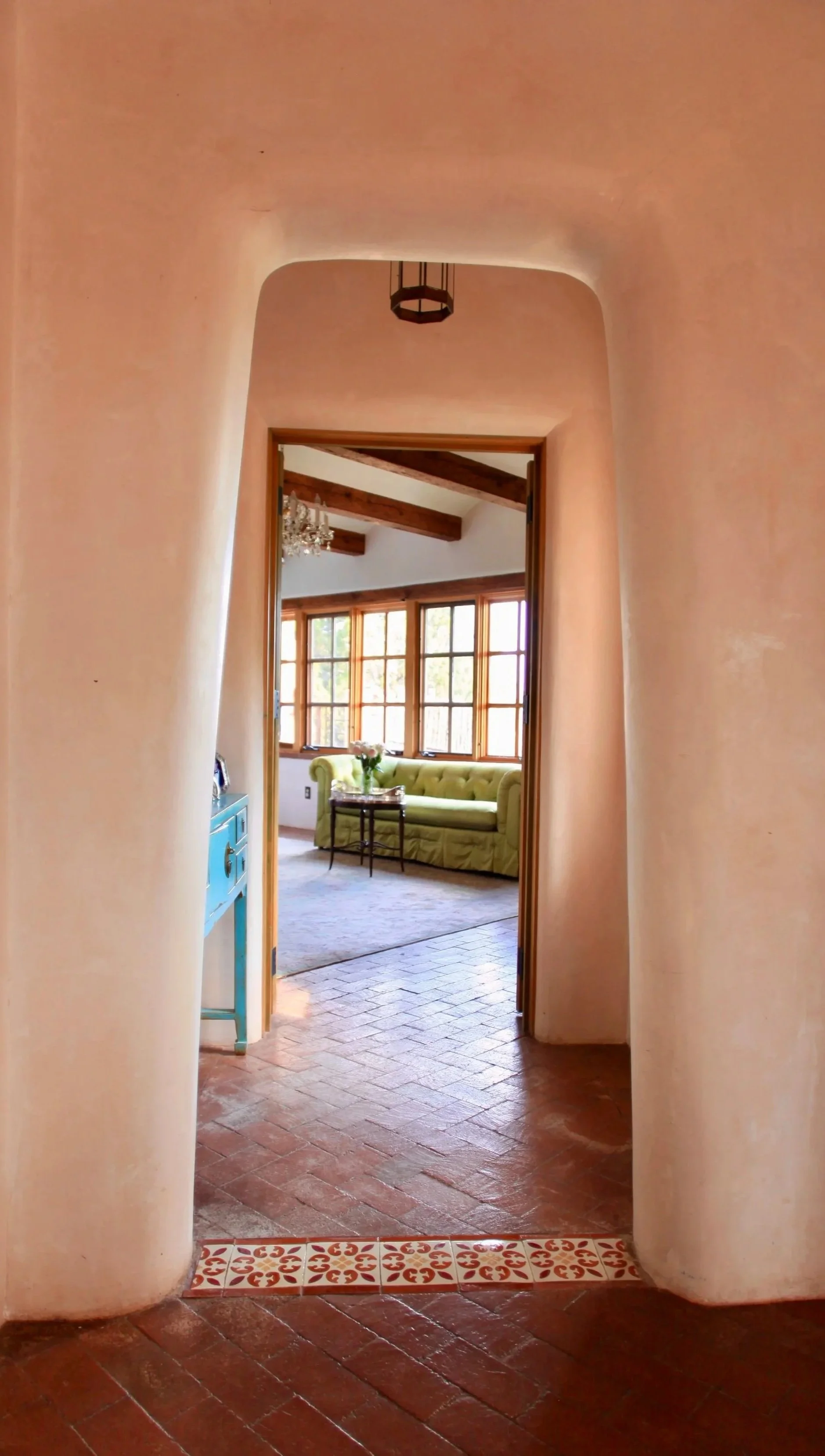 An indoor view through an archway shows a living room with large windows, a green sofa, a side table with vase of flowers, and wooden ceiling beams.