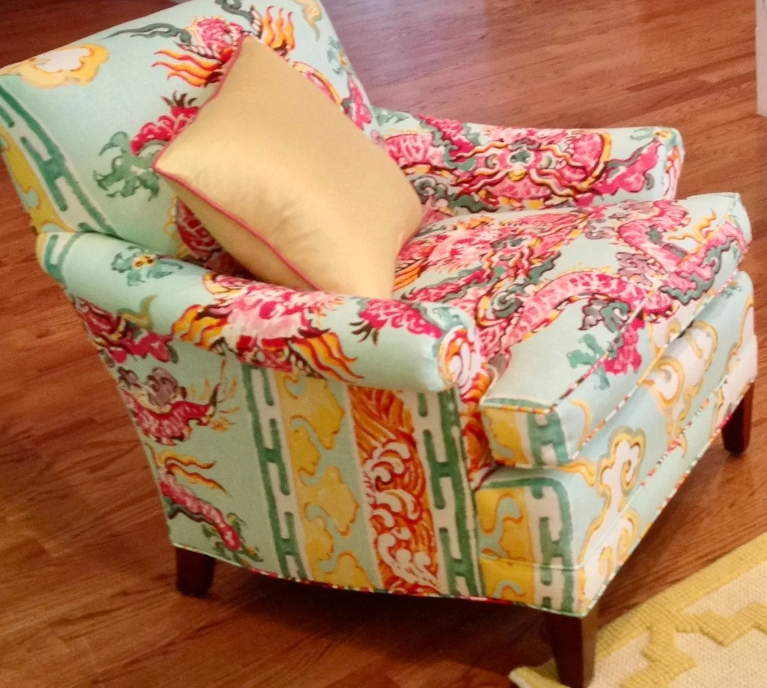 Colorful armchair with intricate floral patterns in green, yellow, pink, and orange, with a light yellow pillow on it, placed on a wooden floor.