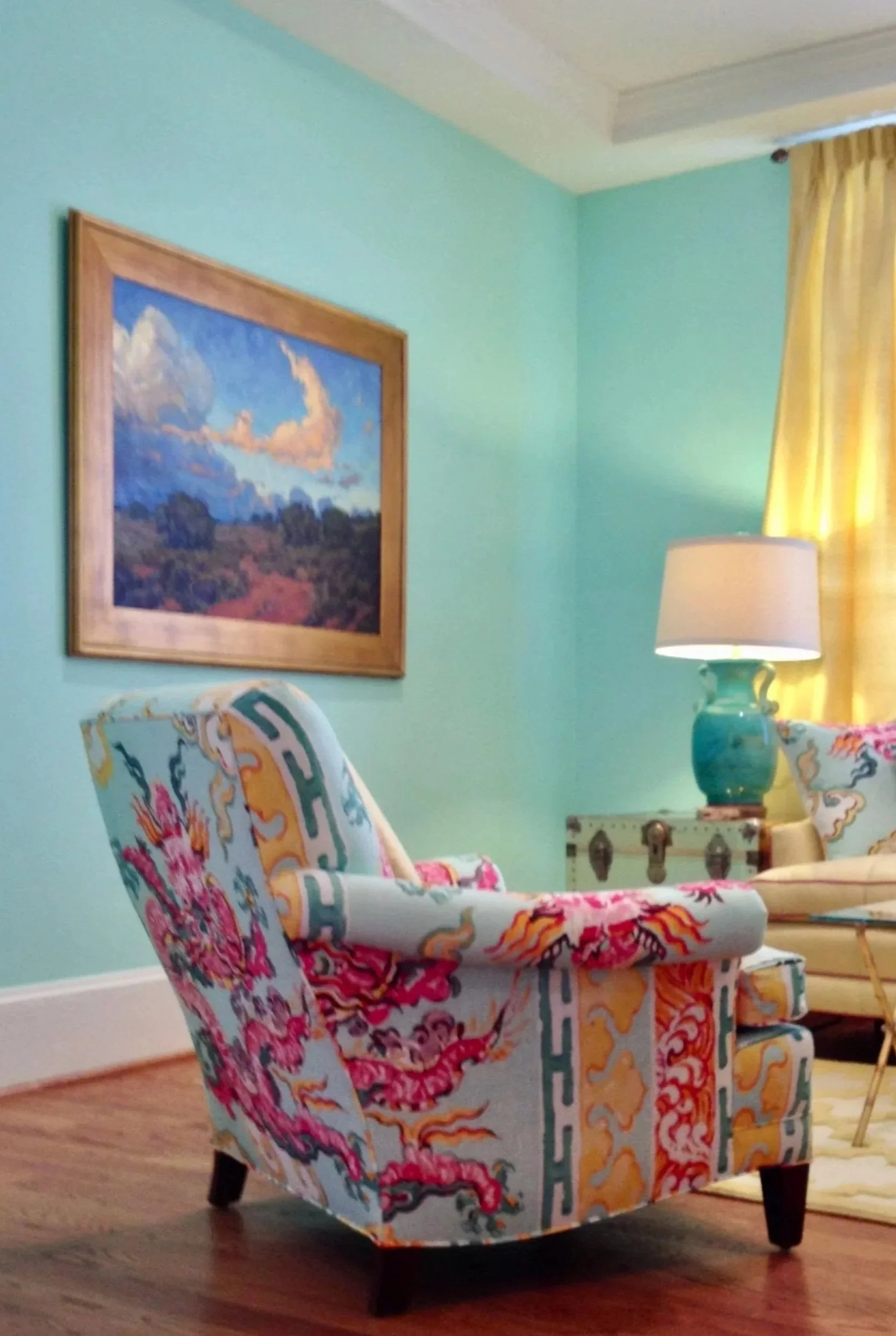 Living room with light blue walls, a colorful upholstered armchair with dragon designs, a side table with a turquoise lamp, a painting of a landscape with clouds, a window with yellow curtains, and hardwood flooring.