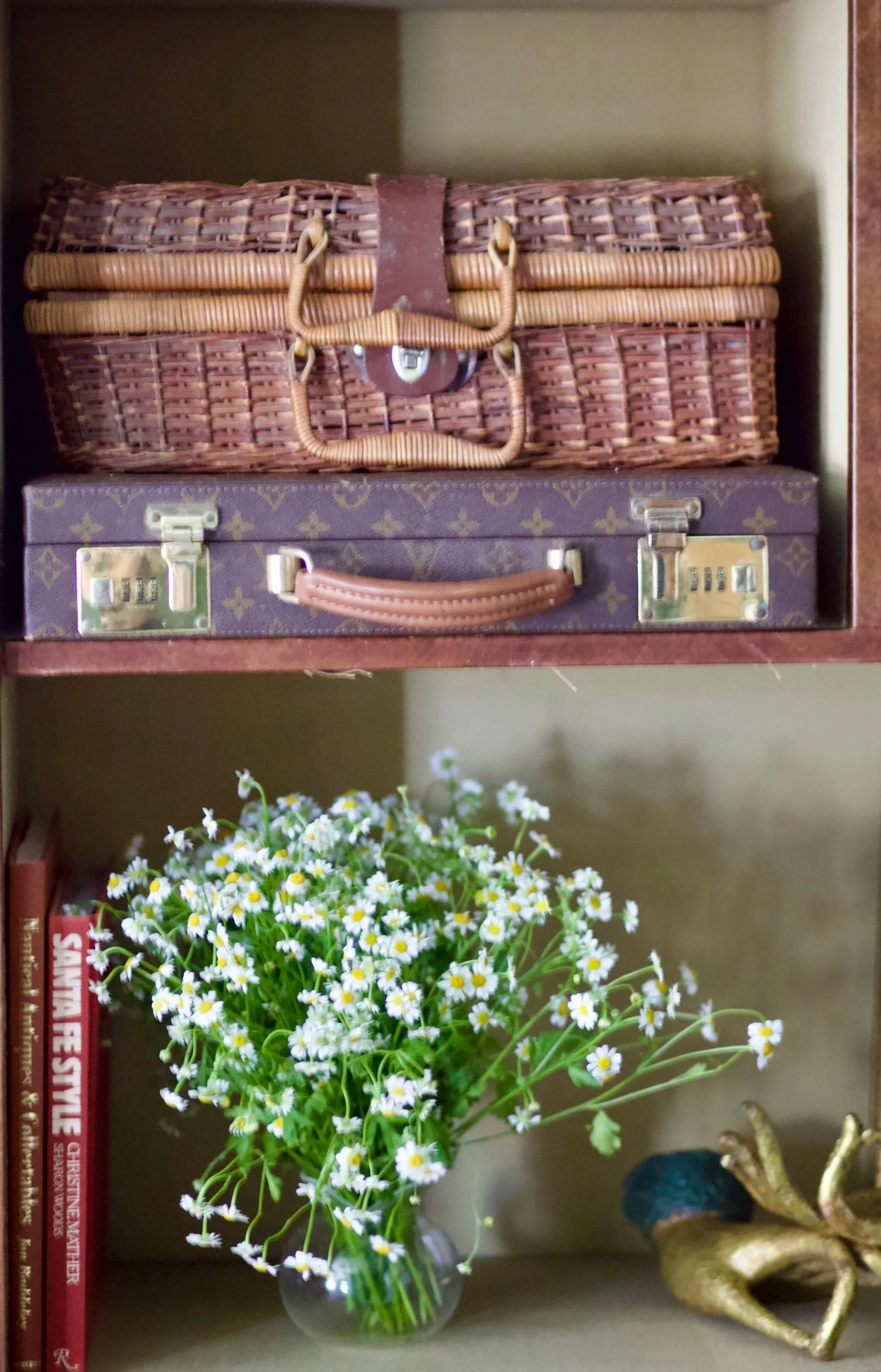 Shelf with two vintage suitcases and a vase of daisies, with books and a decorative object nearby.