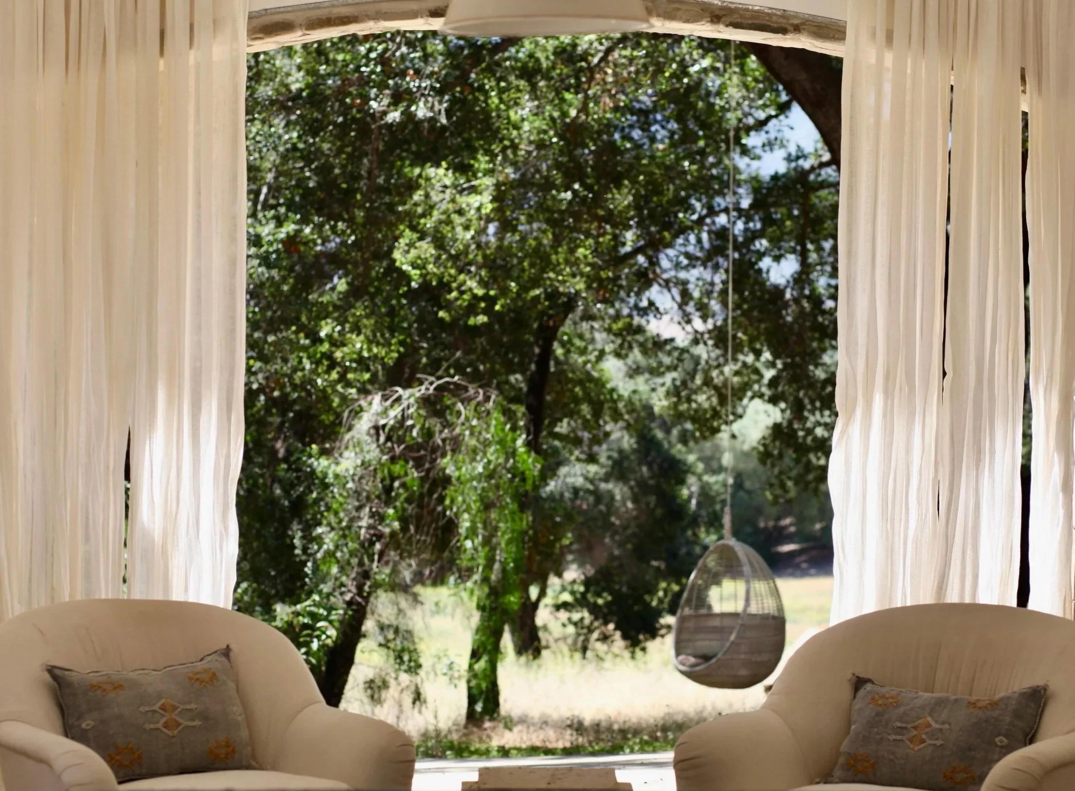 Living room with large window looking out to trees, beige armchairs with patterned pillows, hanging swing chair outside, and white curtains.