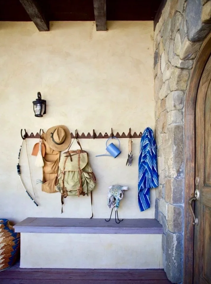 Entryway with coat hooks holding a tan hat, a backpack, a blue watering can, a pair of goggles, and a blue striped towel. A sheep skull decorates the wall below the hooks. A lantern is mounted on the wall above. There is a stone wall and wooden door 