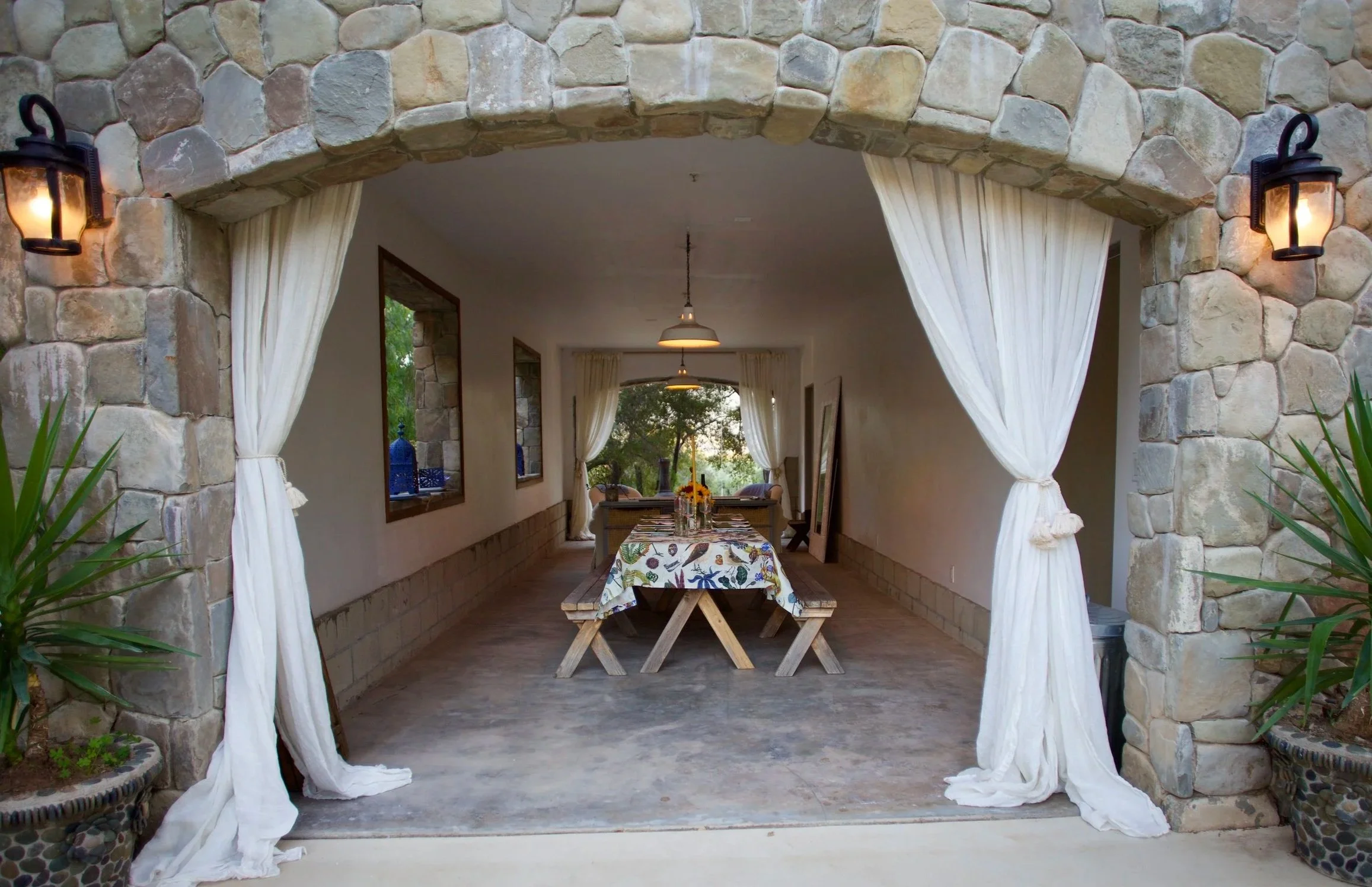 View through an arched stone entrance into a dining area with a wooden table, floral tablecloth, benches, hanging lights, white curtains, and large windows with curtains showing outdoor trees.
