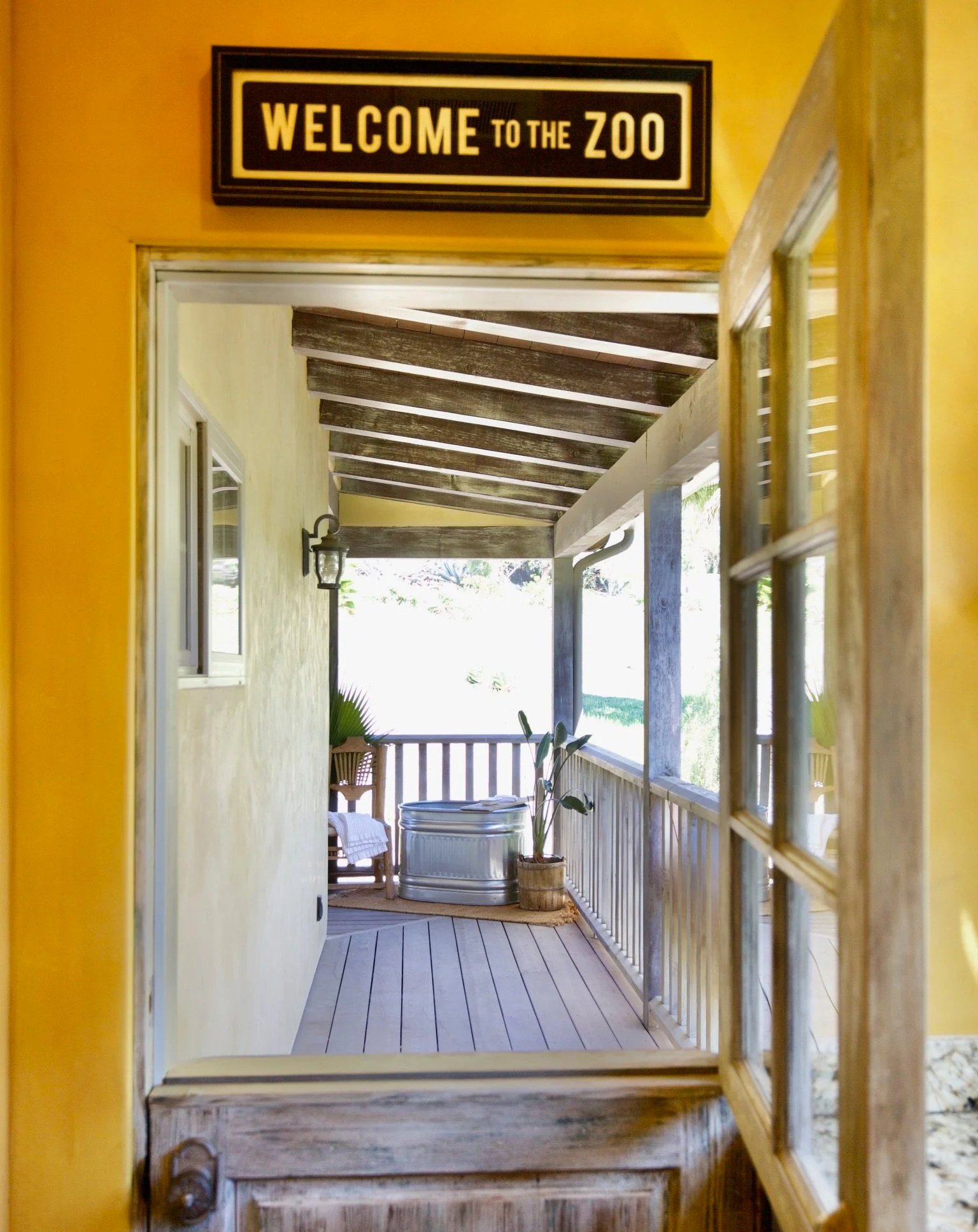 View through a wooden door into a porch with a yellow wall, potted plant, and outdoor furniture, with a welcoming sign that says 'Welcome to the Zoo' above the doorframe.