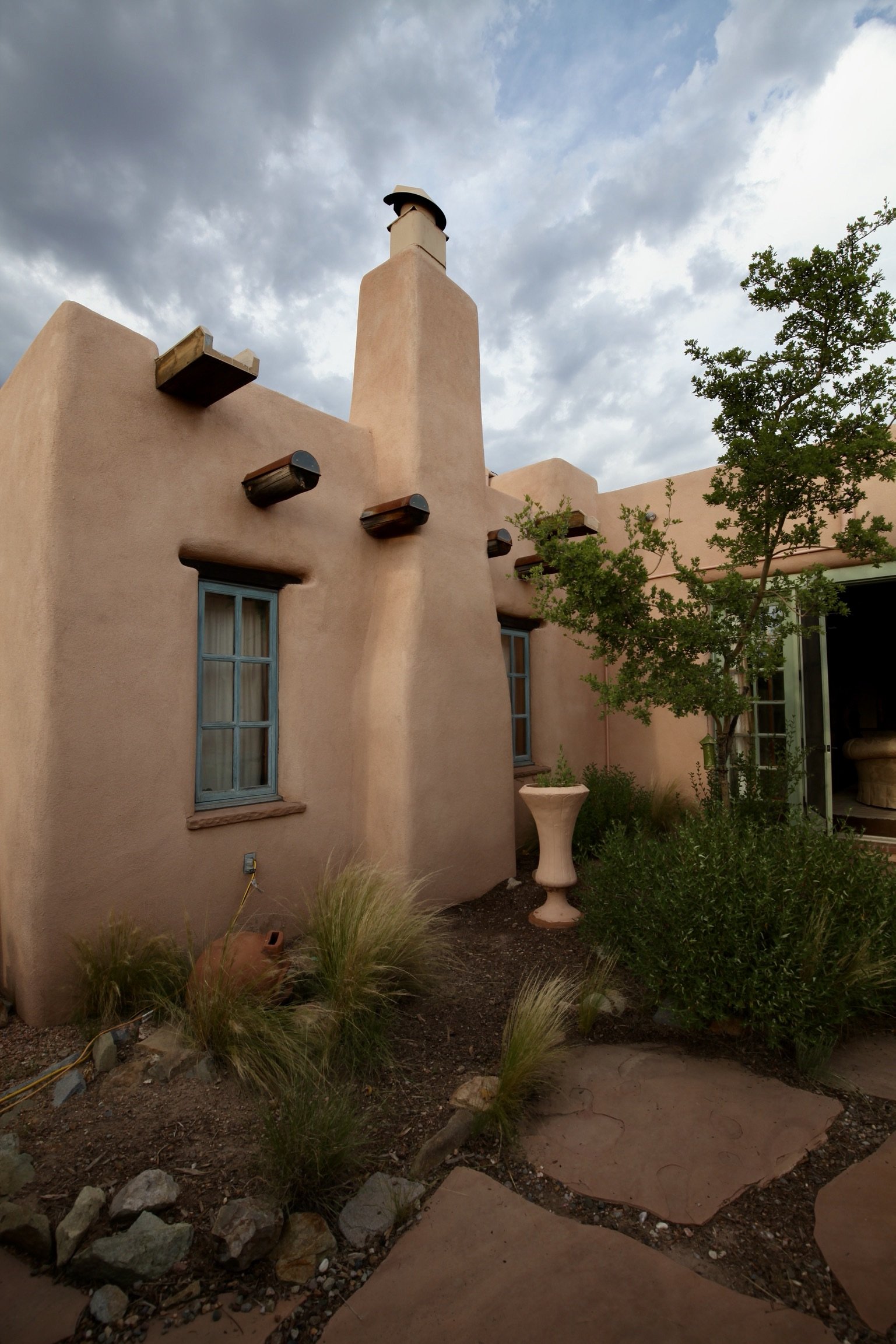 Adobe-style house with stucco walls, small windows with blue trim, chimney, and a garden with plants and a large urn, under a cloudy sky.