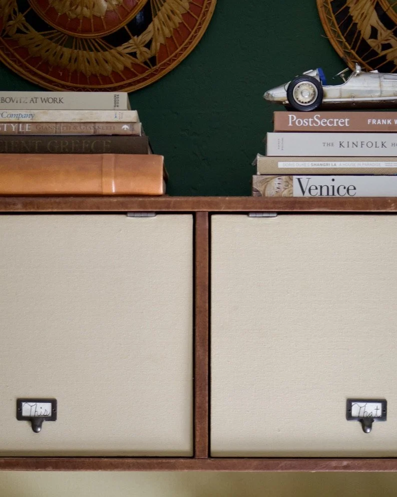 A vintage wooden cabinet with two beige fabric drawers labeled "This" and "That". The top of the cabinet holds a stack of books, a model car, and decorative wall hangings.